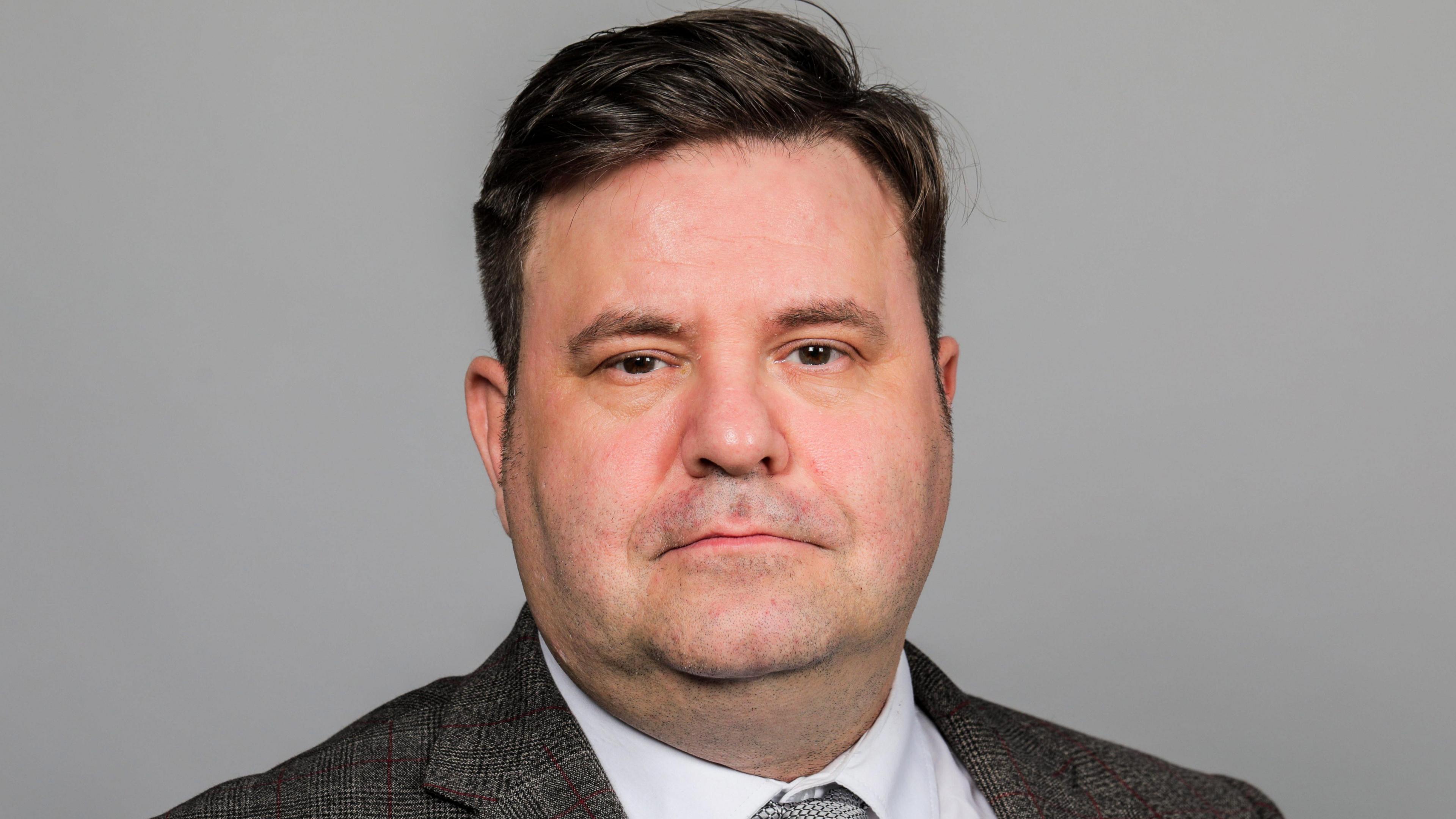 A head and shoulders shot of Alec Brown who has dark hair and is wearing a grey suit and tie.