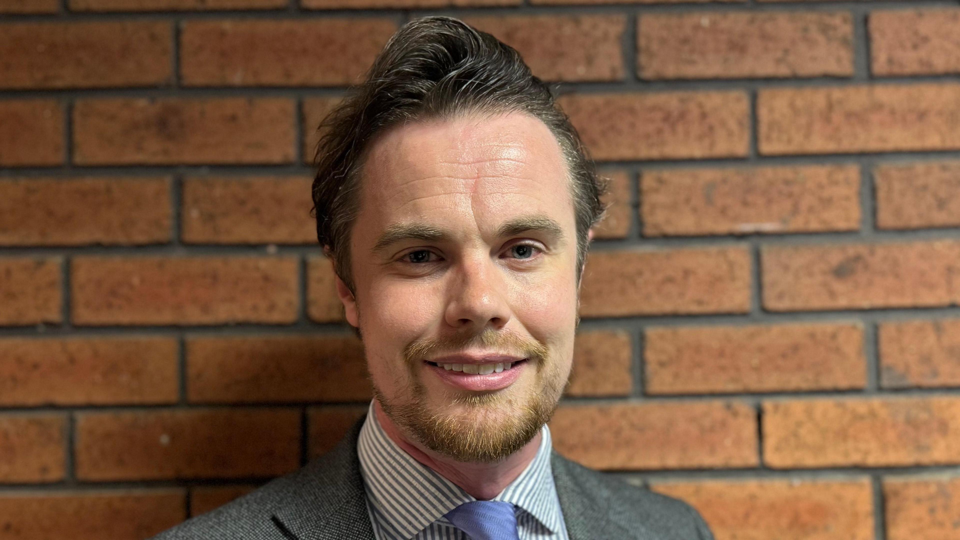 A general view of Philip Faircloth-Mutton. He smiles at the camera while standing in front of a red brick wall. He has short dark hair and a beard. He wears a grey suit, a shirt and blue tie.