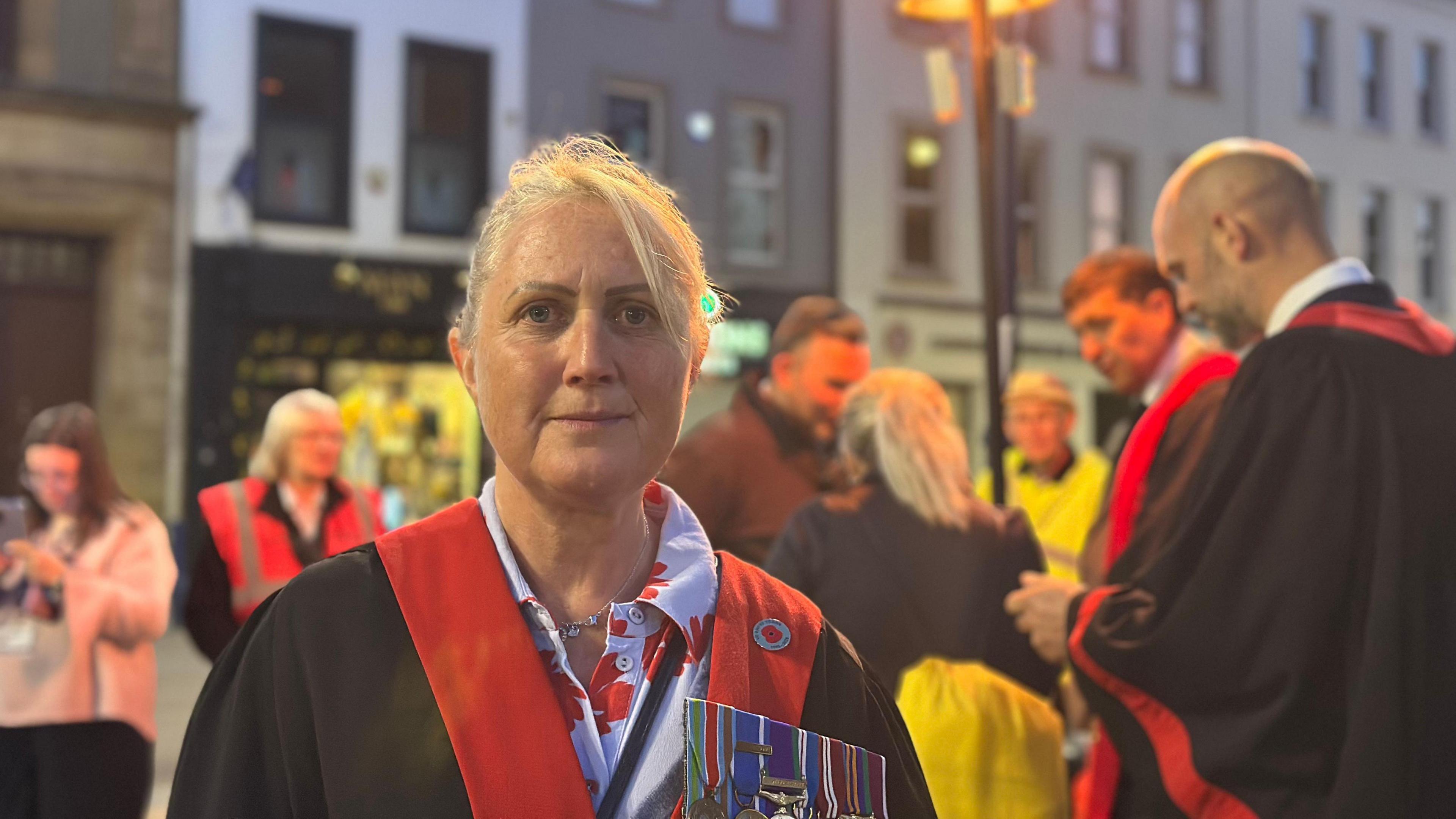 A woman with blond hair, wearing a black and red robe. She is looking at the camera. There is a group of people standing in the background.