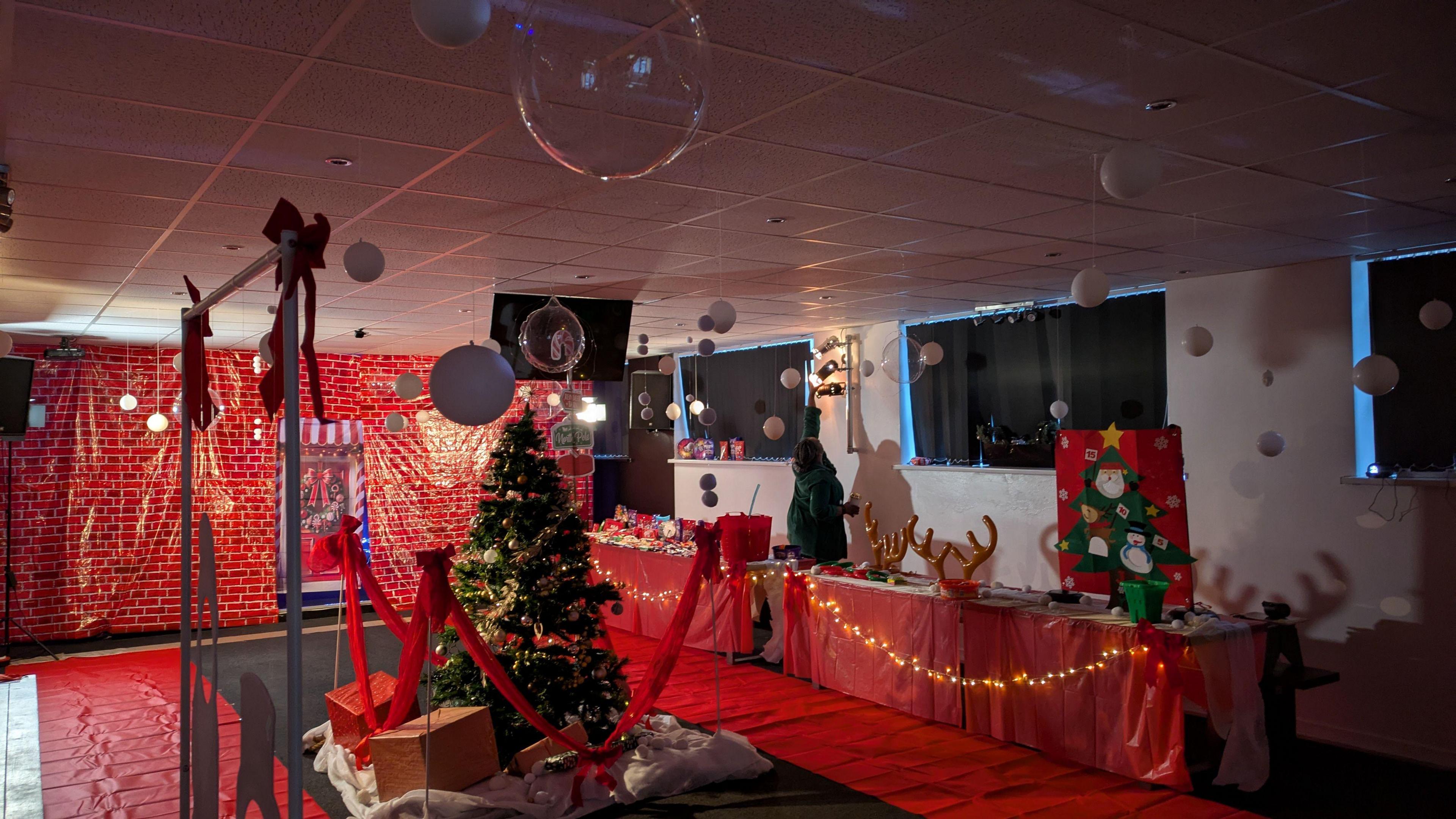 A church hall covered in red festive decorations. A sheet covers the back wall which is patterned with red bricks and a festive doorway. White and clear balloons hang from the ceiling and a Christmas tree is covered in baubles in the middle of the room. Tables are covered in red tablecloths, fairy lights, and Christmas decorations.