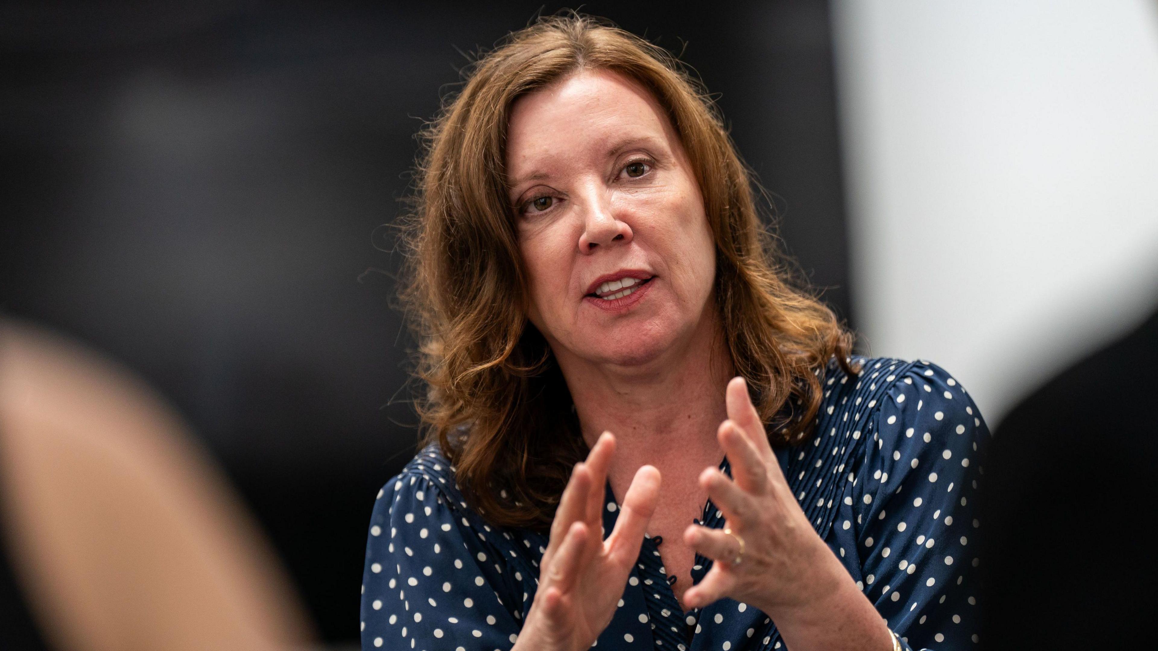 Rachel De Souza giving a talk. The background is blurred. She is wearing a blue and white top and has her hands in front of her. 