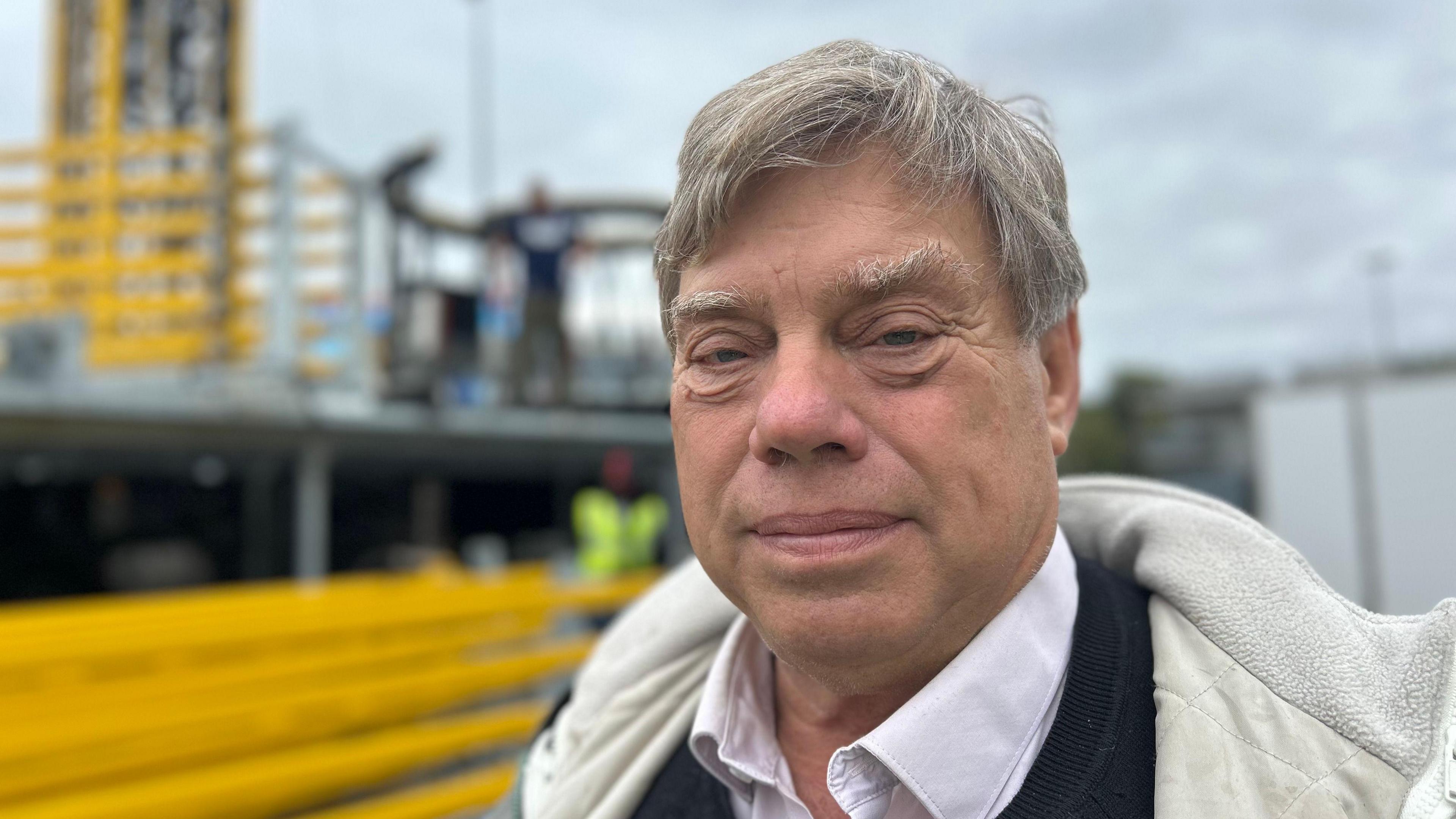 A man wearing a white shirt with a black jumper over it, he has blue eyes white hair and bushy eyebrows, he's smiling and is stood next to an 82m ride, he looks glad to be at the fair and is stood close to the camera.