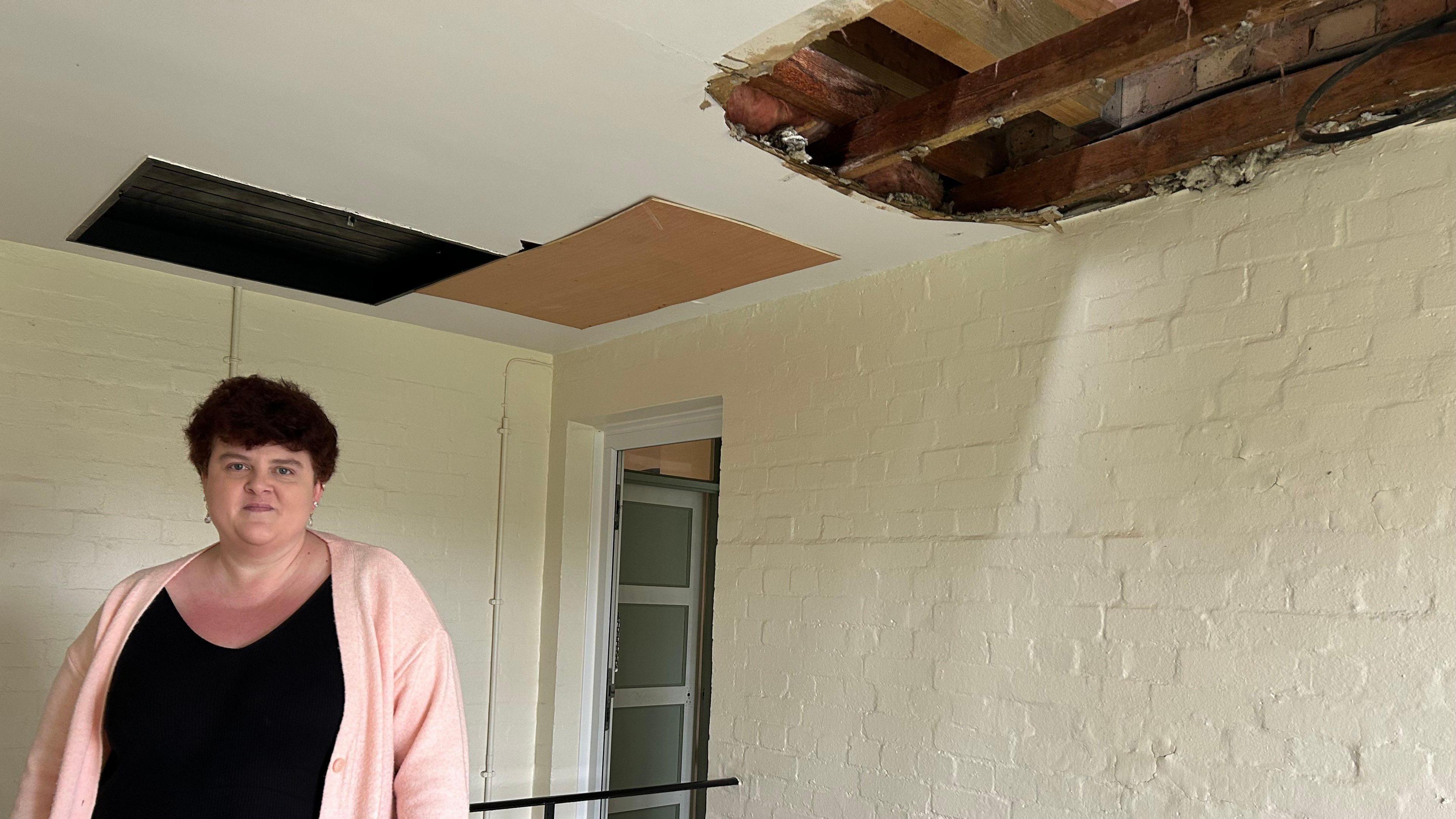 Sarah Carson standing on the left wearing a peach cardigan and black top. On the ceiling to the right is the hole exposing parts of wood and brick wall.