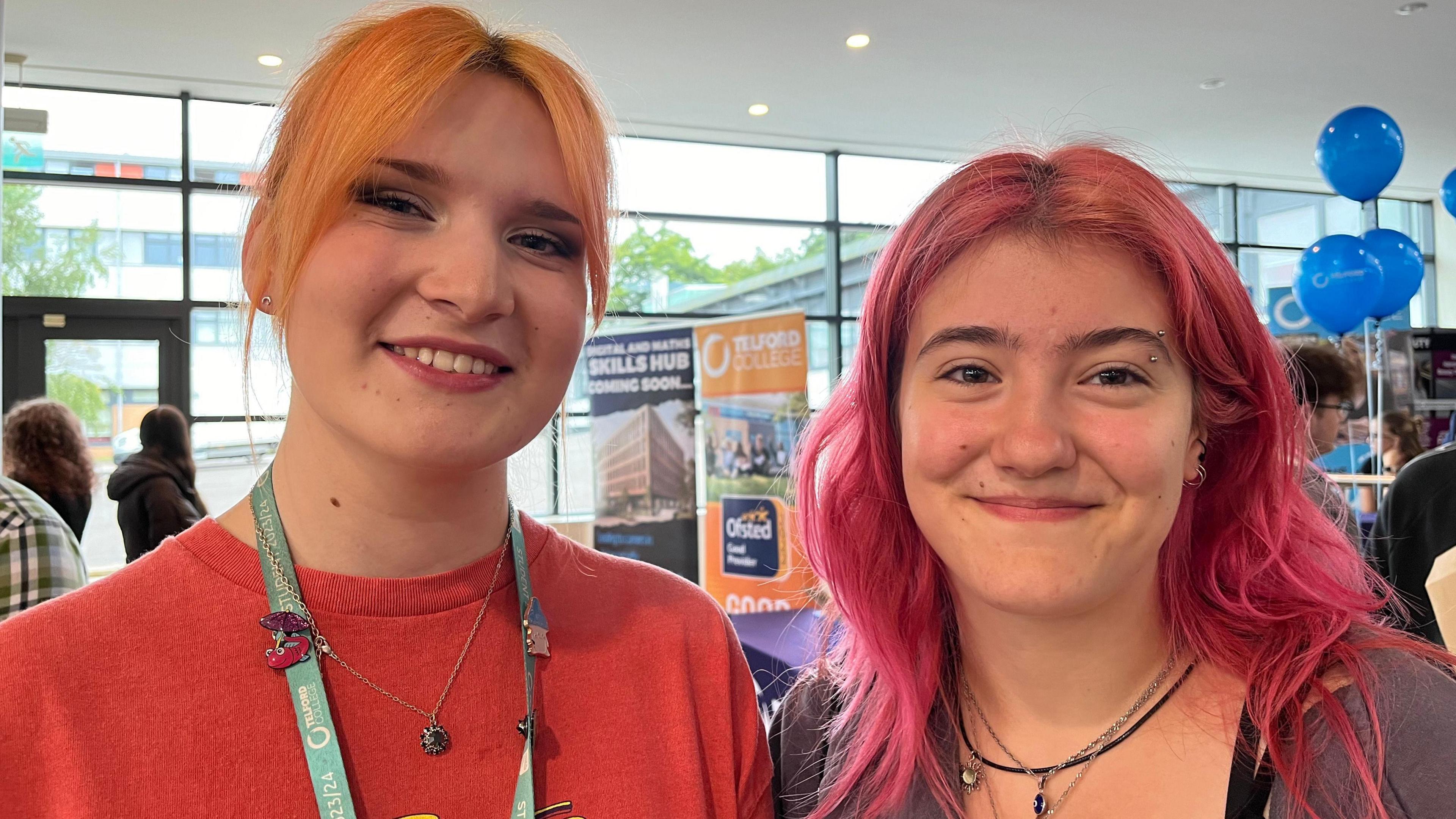 Maddie Bethwaite is standing on the left-hand side. She is wearing a red jumper and a light blue lanyard around her neck. She has orange hair. Maya Newton is wearing a purple top. She has pink hair and has her eyebrow pierced.