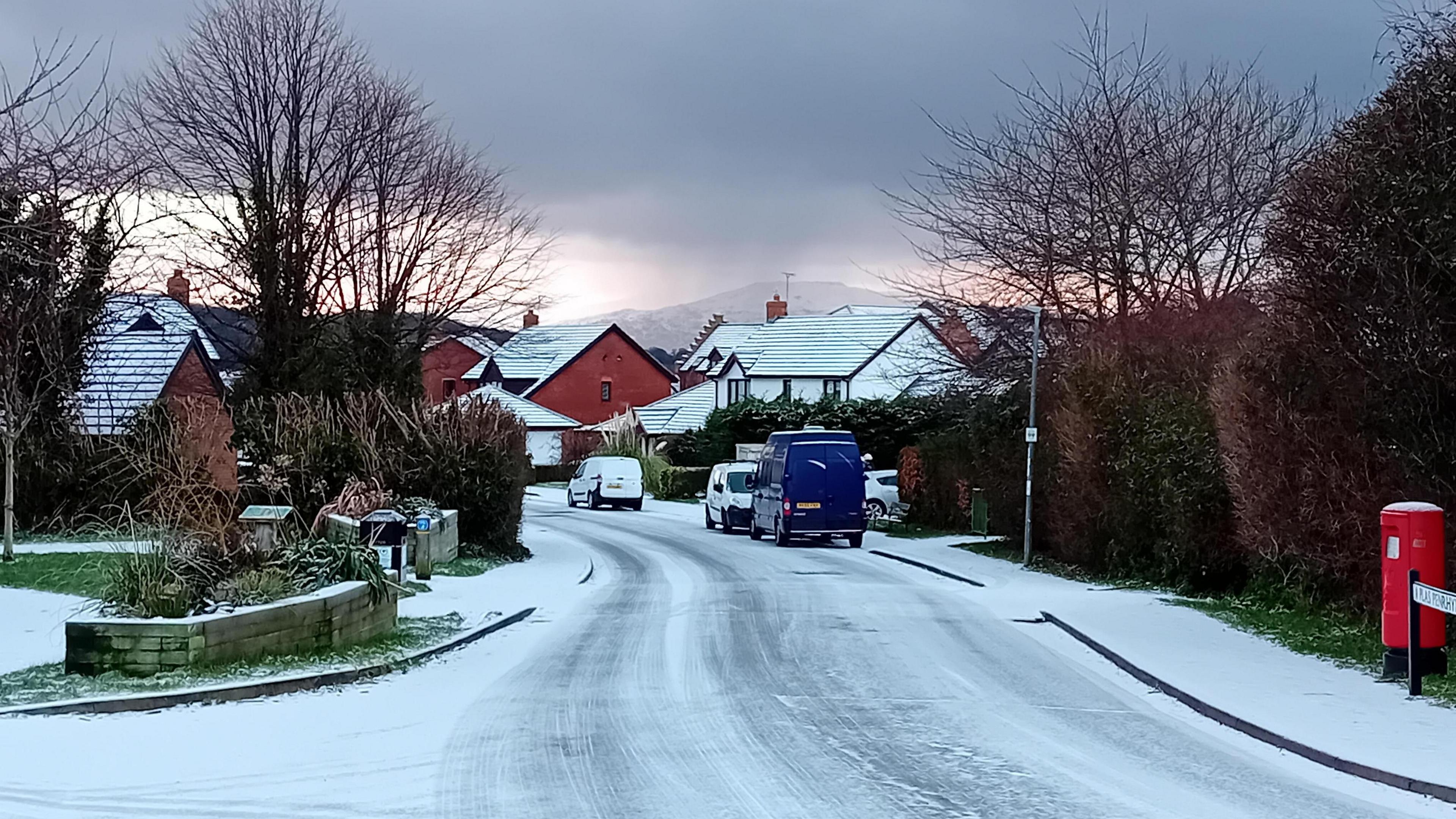 Road in residential area covered in thin layer of snow and ice with cars and van parked along the road