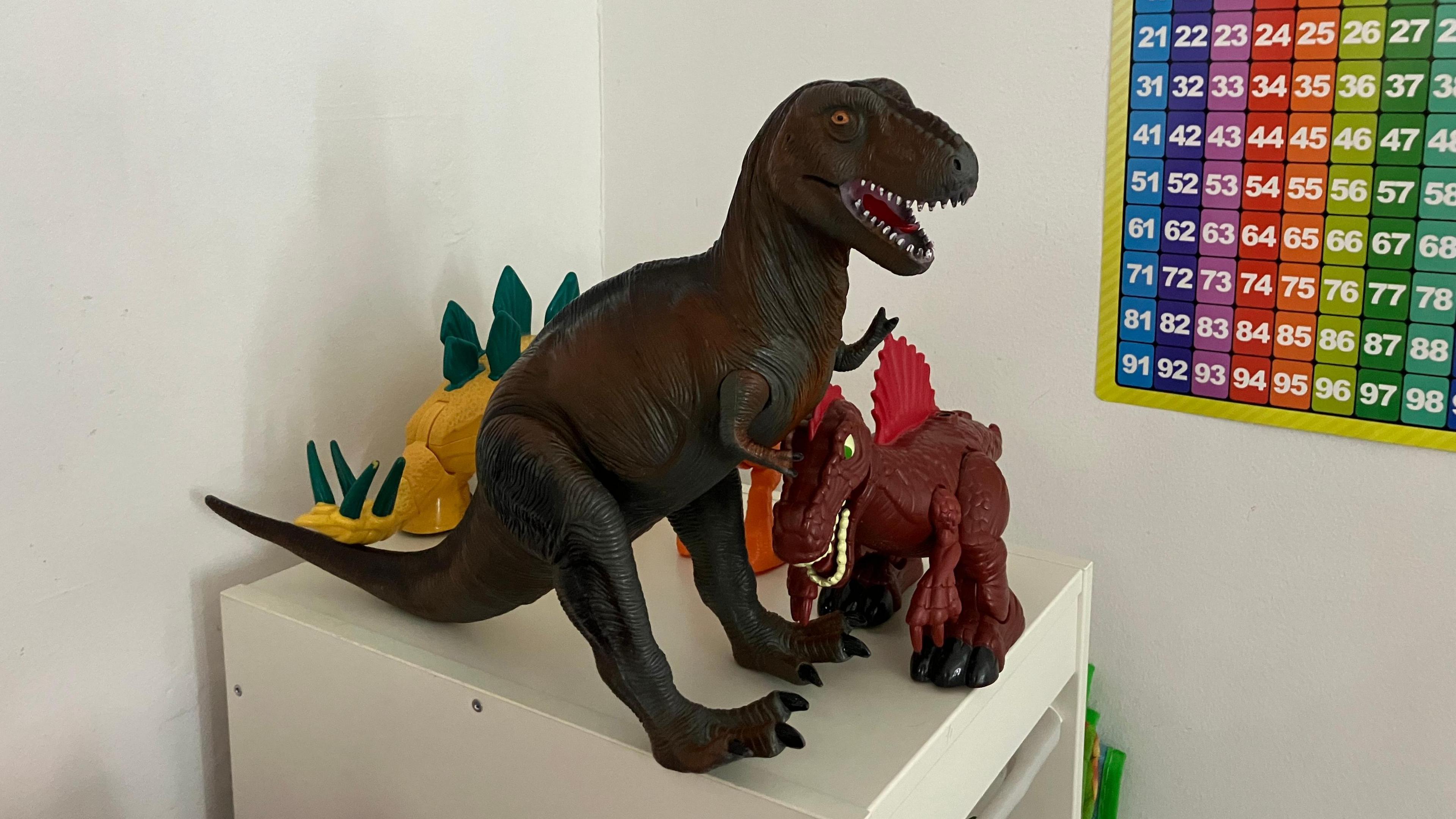 Three toy dinosaurs perched on the top of a white cabinet. On the right of the image is a colourful counting chart.