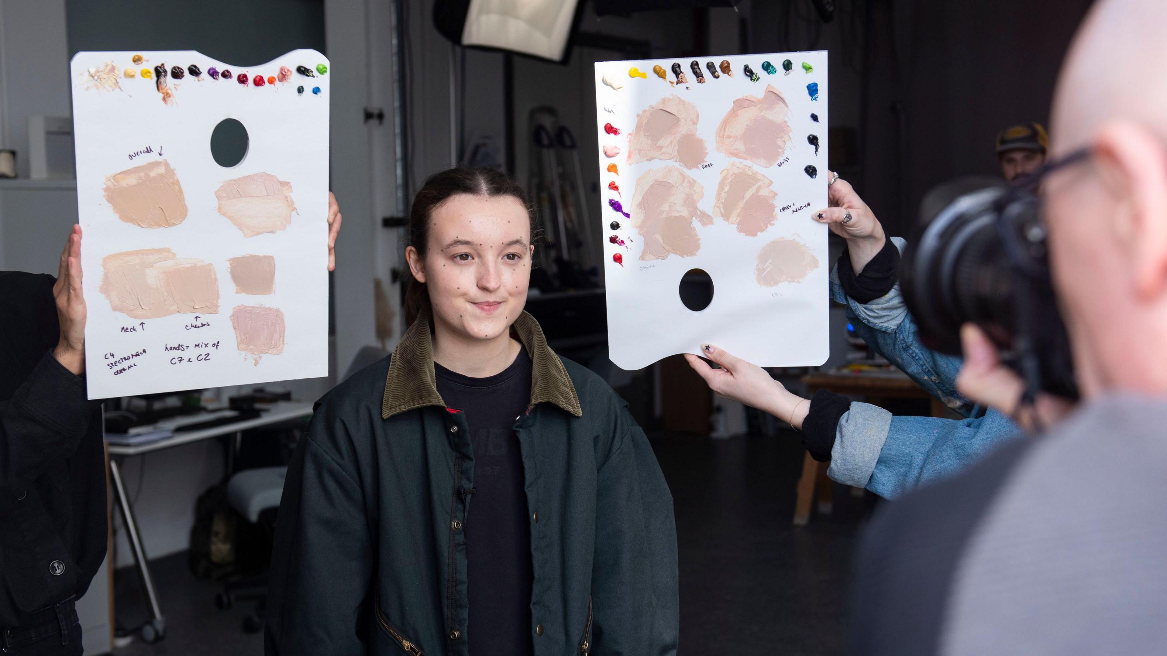 Bella Ramsey with dots on their face and two pieces of canvas are being held on either side of them during the measuring up process for their first figure.