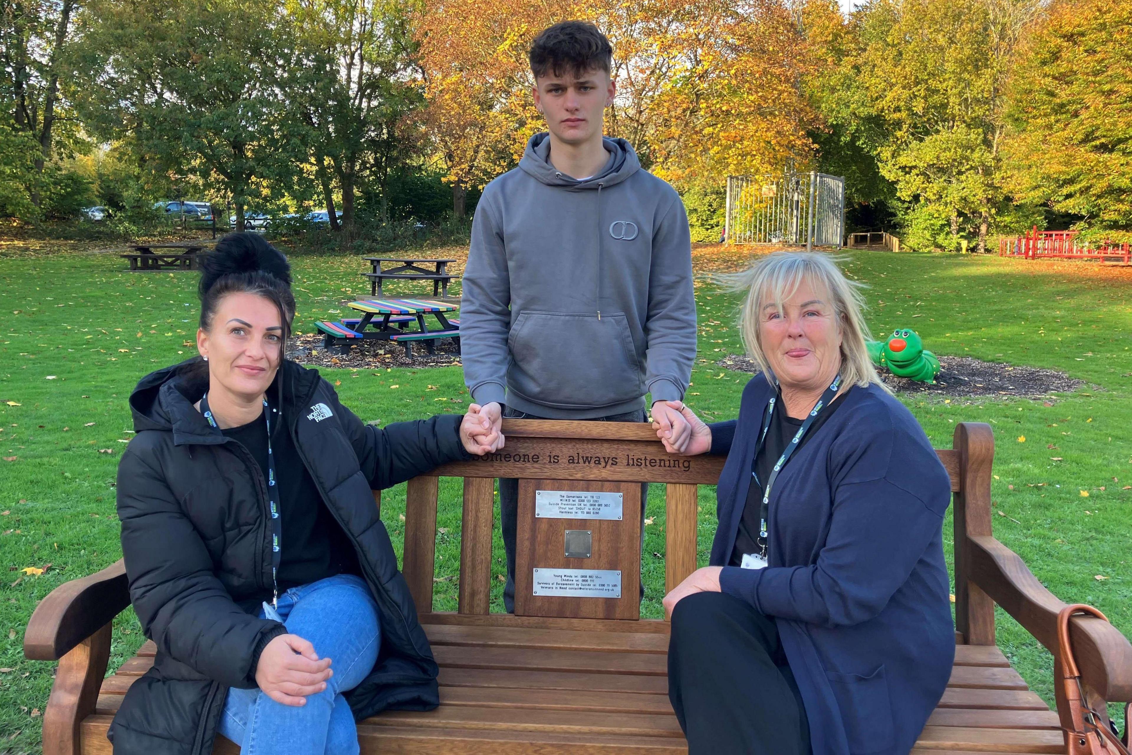 Woman with dark hair, dark jacket and jeans with woman with blonde hair, dark jacket and dark trousers. Boy with short brown hair and grey hoody. The women are sat on a bench and the boy is stood behind it. There is grass.