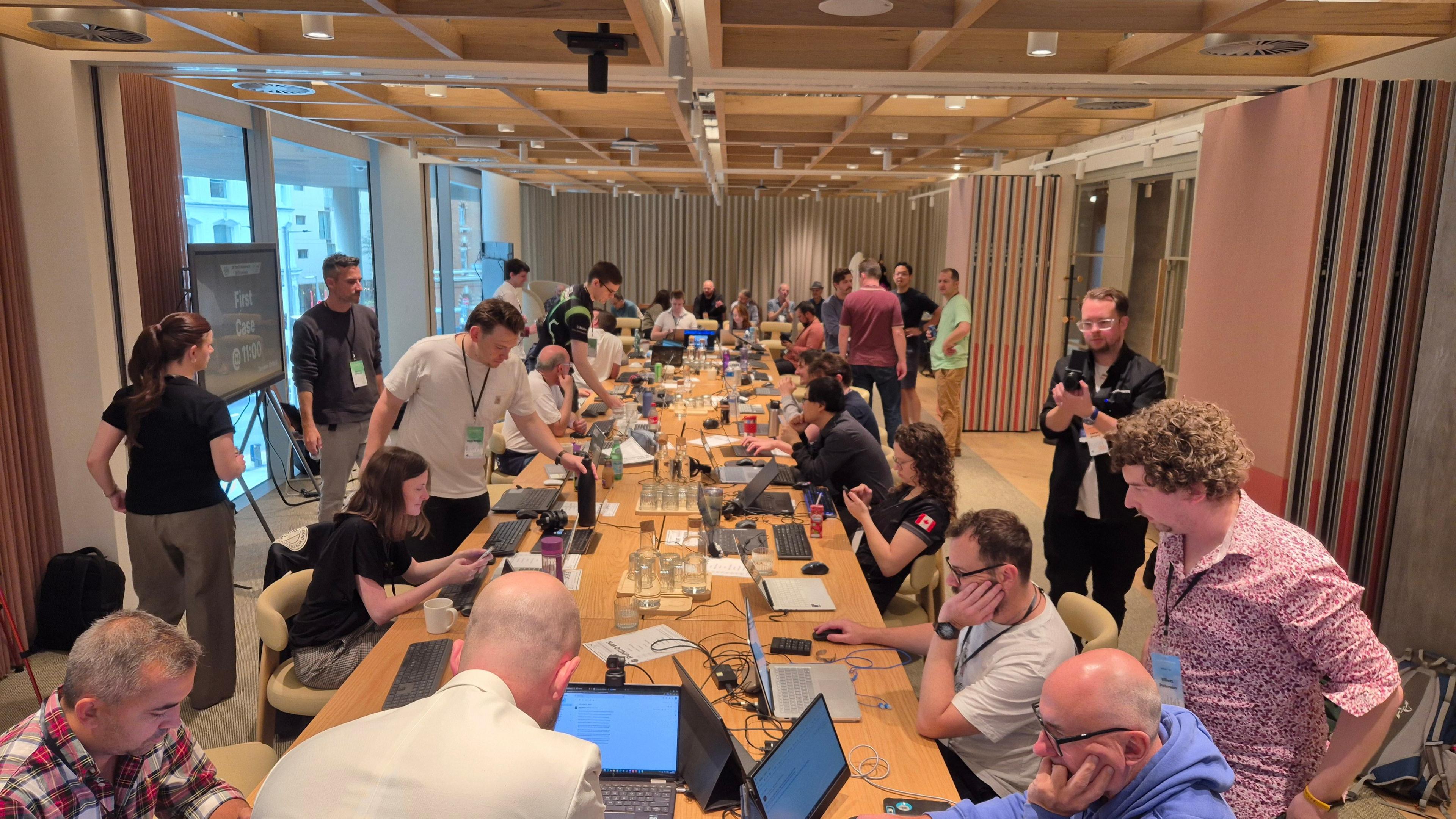 UK championship competitors sat around a very long table in a room full of people. The scene looks very buzzy and lots of people are sat in front of computers.