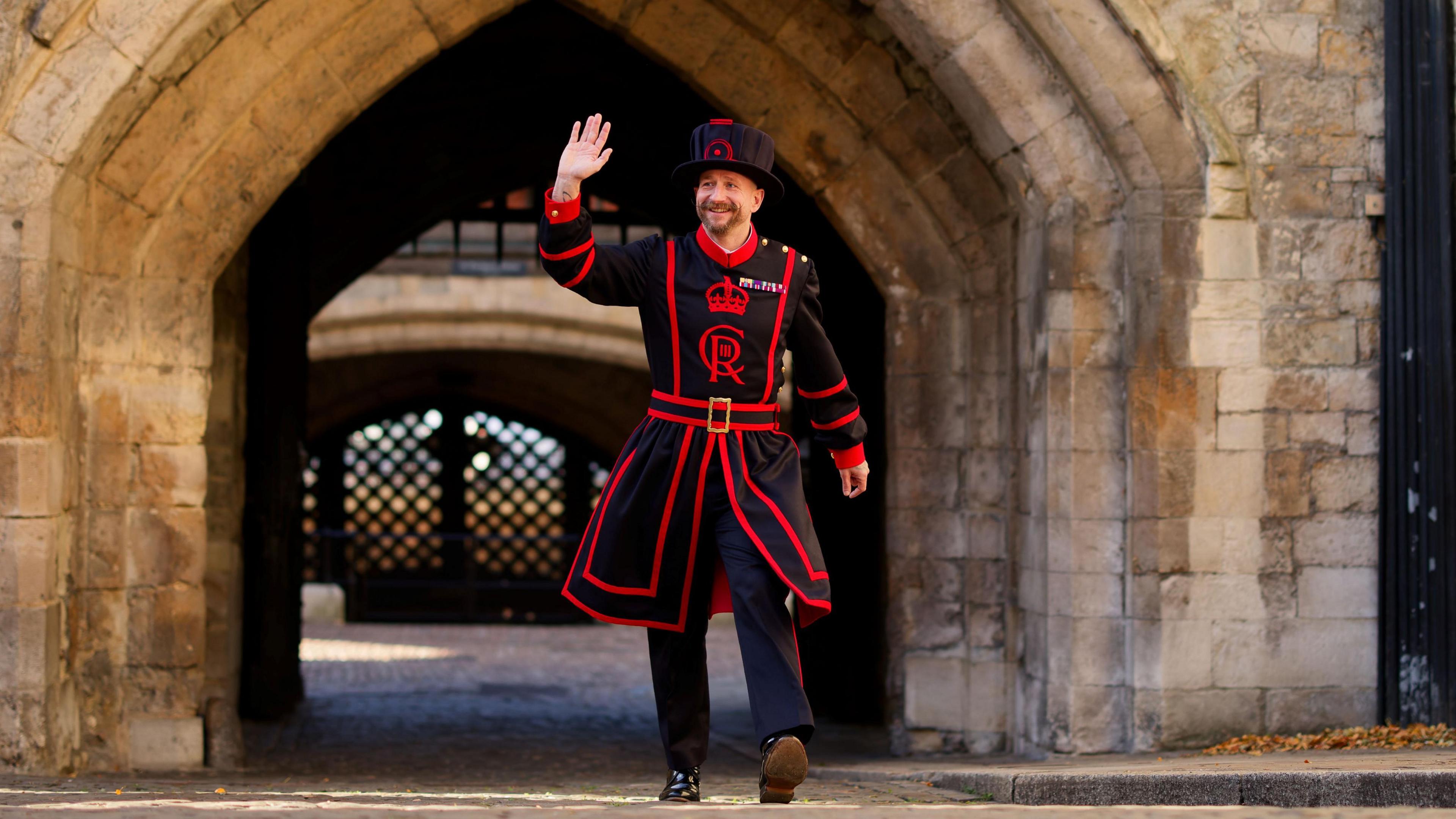 A man in Yeoman Warder uniform - a black cape with red outlines, black trousers, and a black cap. He is waving and walking through an archway. 