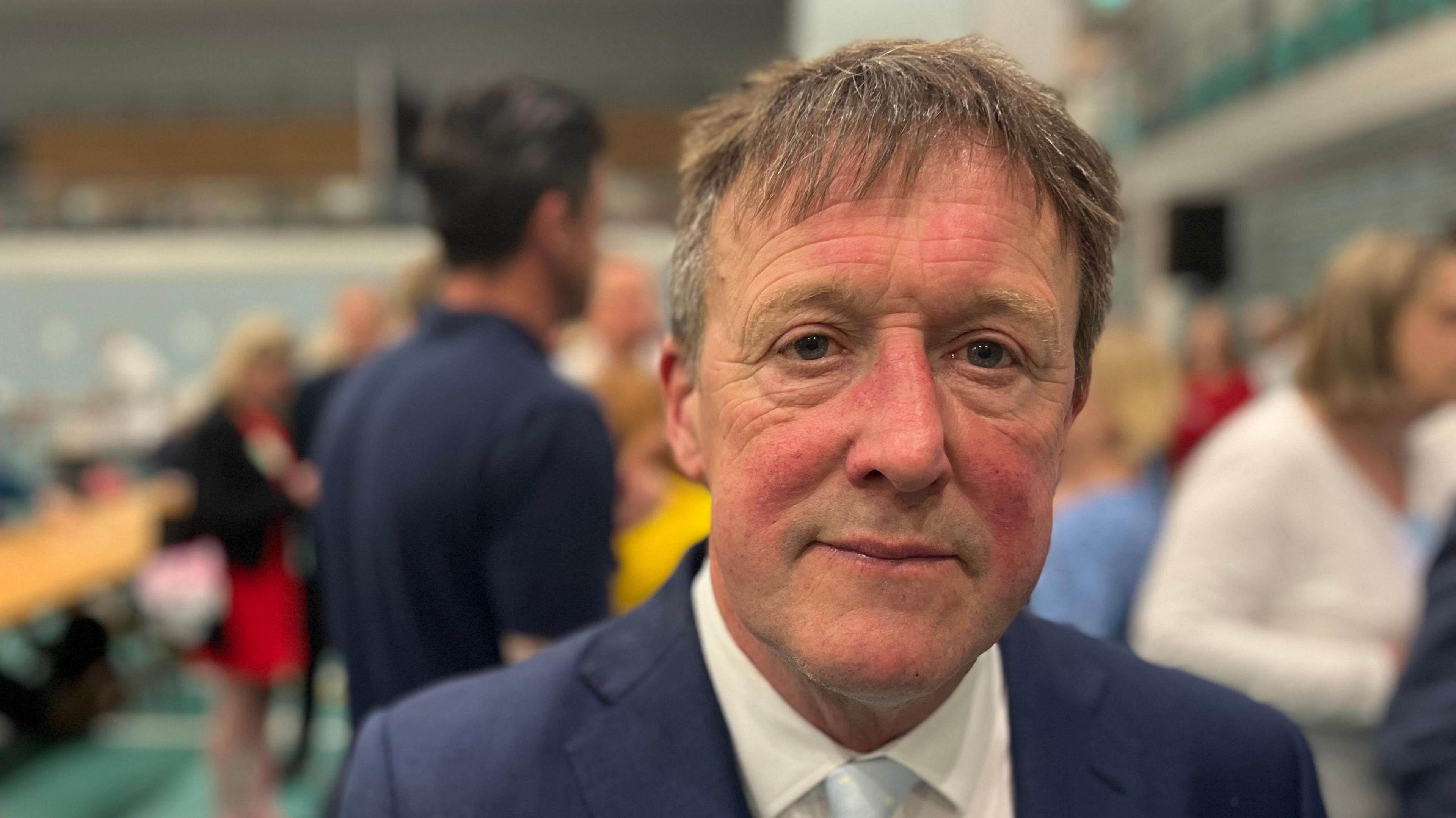 A picture of Neil looking at the camera. He is wearing a blue suit with a white shirt and light blue tie.