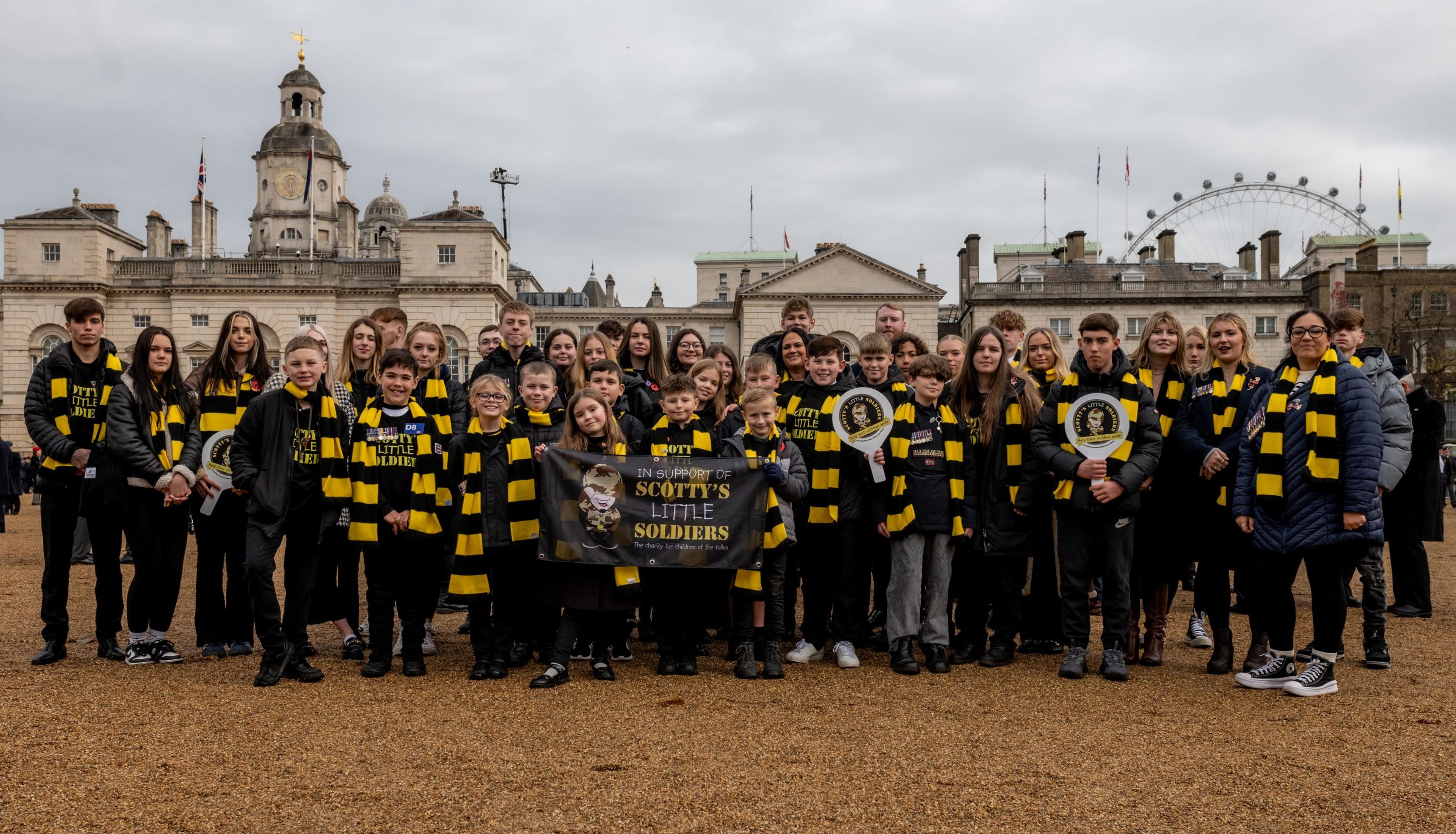 A large group of young people, and several adults, standing outside a building in London, with the London Eye in the distance. They are all wearing black and yellow scarves. Three people are holding up a banner and two are holding paddles. They are all smiling and looking straight at the camera. 