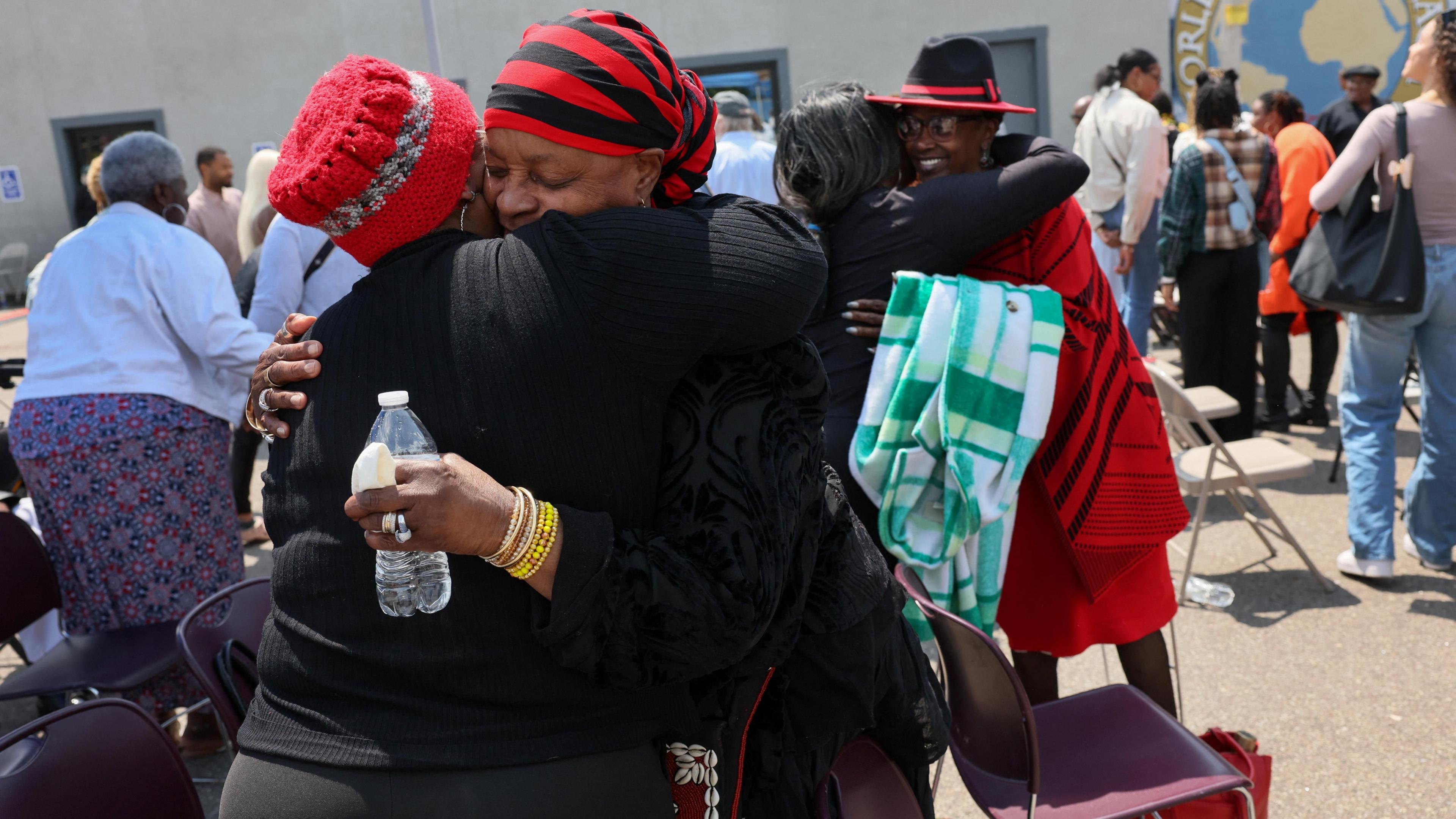 Women dressed in red and black hug in a parking lot next to folding chairs