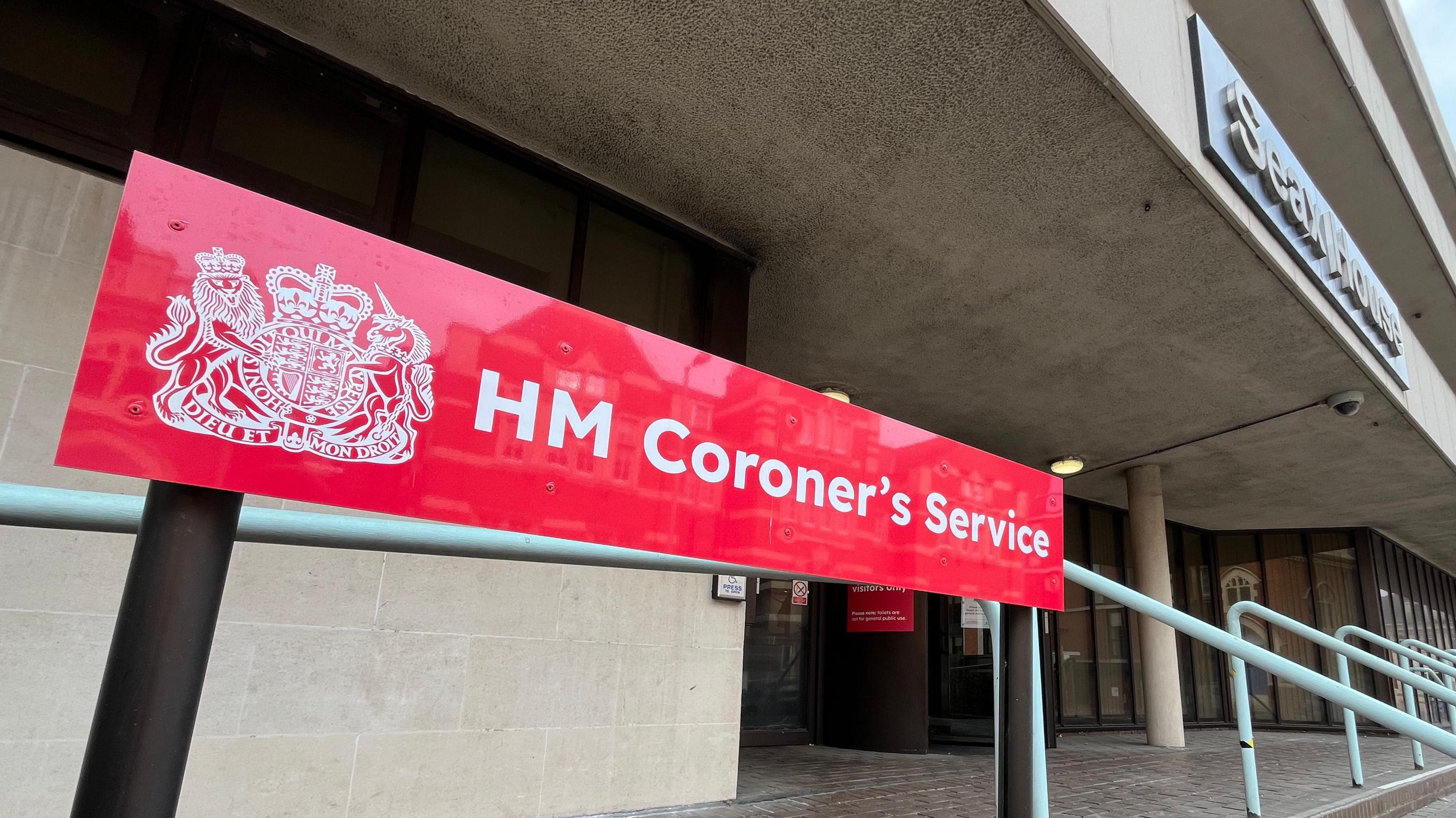 A red sign that says HM Coroner's Service. It is at the entrance to Essex Coroner's Court, which is a cream building that has a black revolving door and brick paving outside.