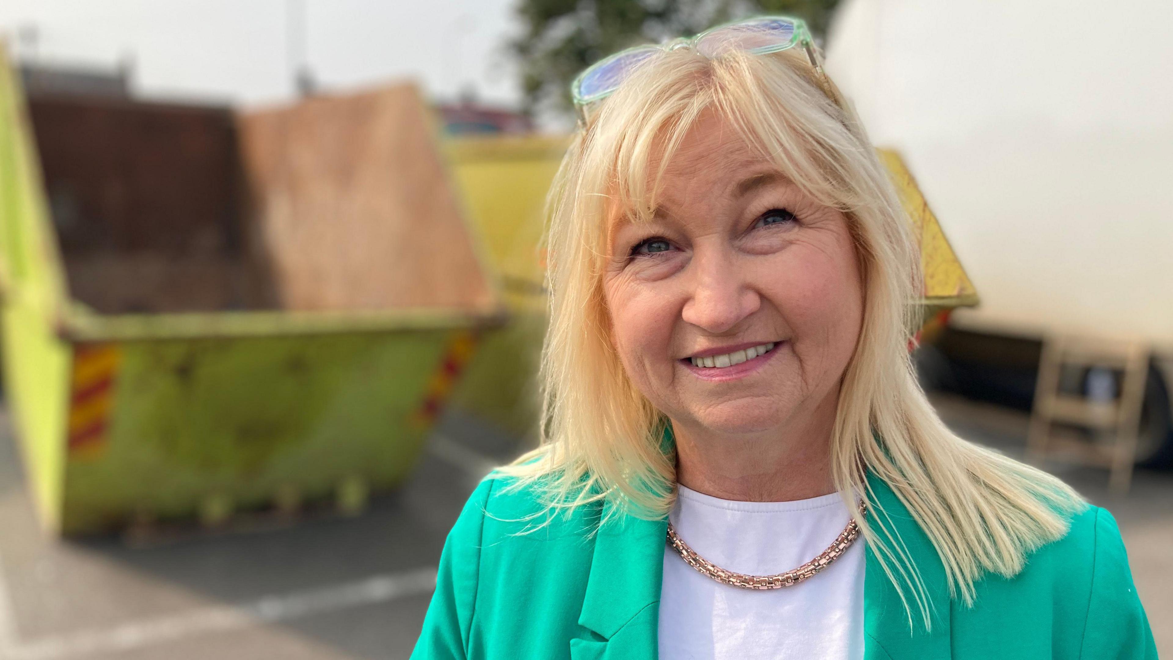 Council leader Anne Dorrian standing in front of a yellow skip wearing a green jacket, white top and gold chain. She has shoulder-length blonde hair and has glasses propped up on her head