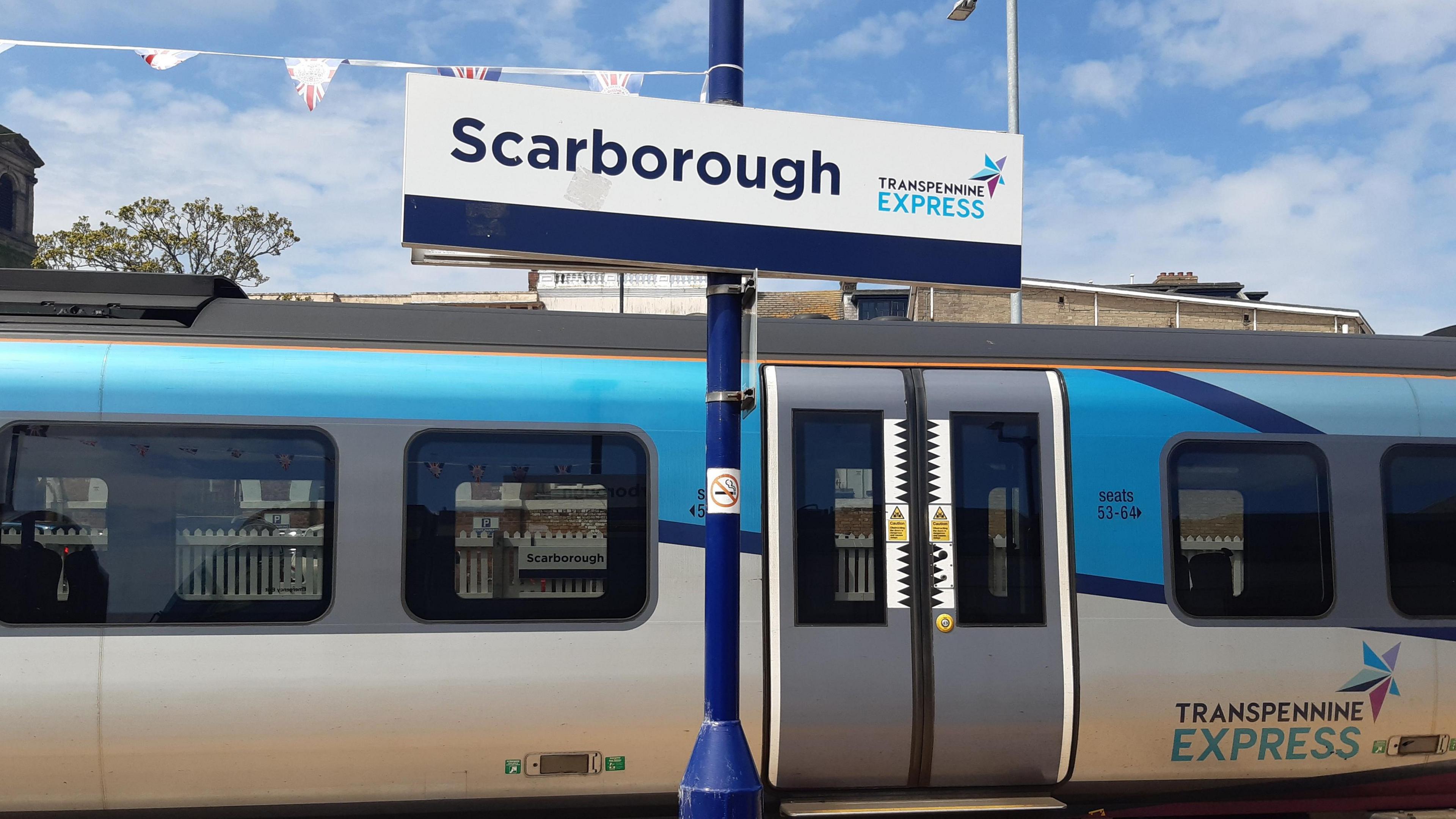 A railway station sign reading Scarborough on a platform with a train in blue and white livery behind it