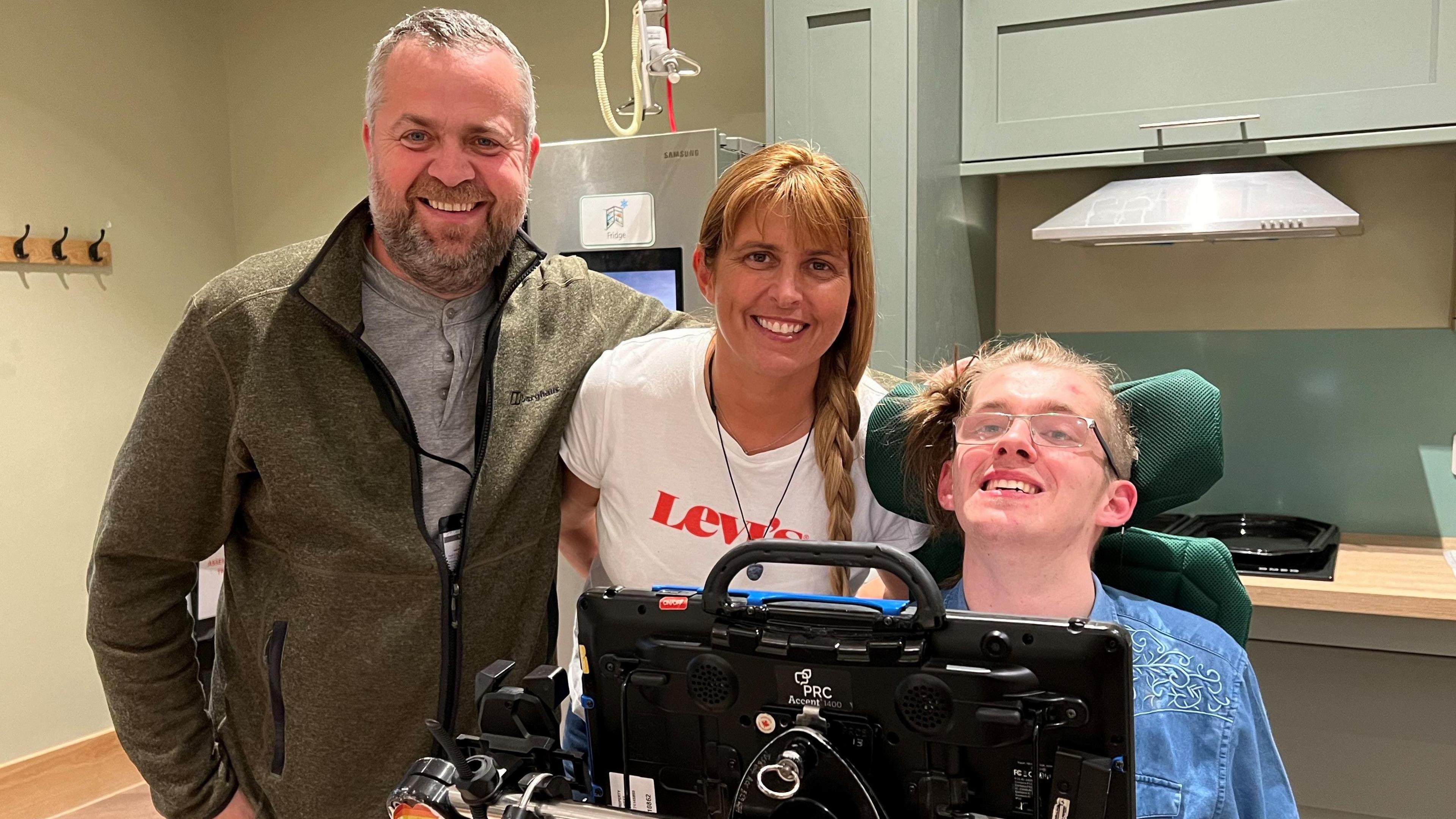 A man in a grey top with a green jacket on, next to a woman with ginger hair in a plait with a white op on, next to a man with a blue top on and in a wheelchair. 