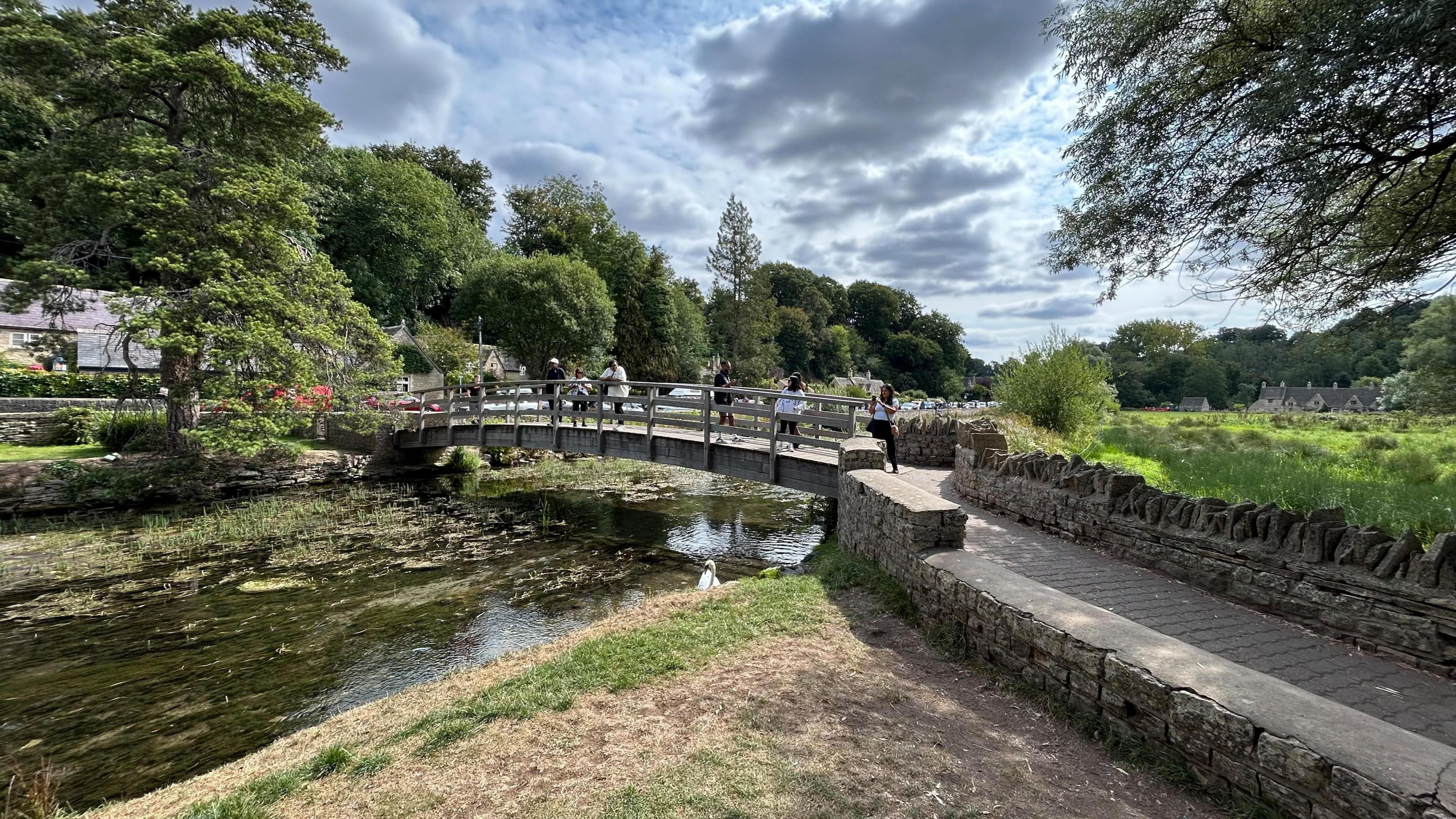 A view of a wooden bridge over a river. There are trees surrounding the scene and people walking across the bridge. It is a sunny day with some cloud.