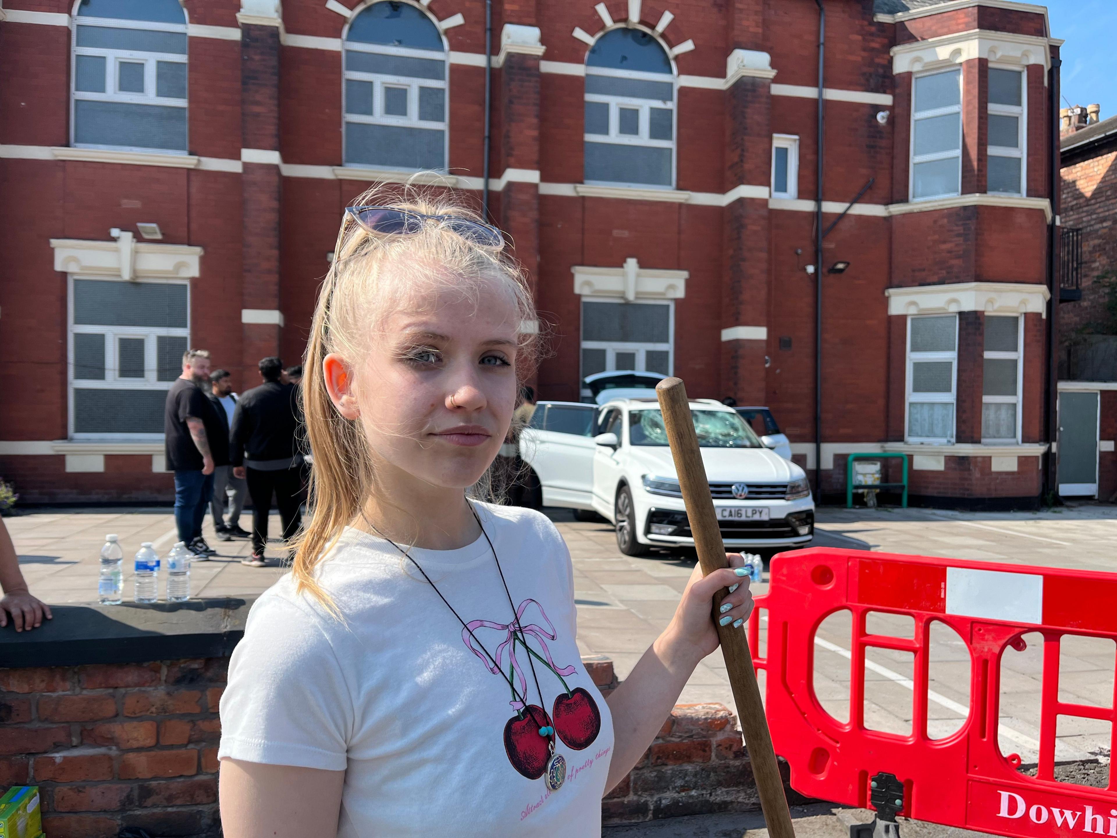 Beatrice looking into the camera and holding the handle of a broom, she is dressed in a white T-shirt and standing outside the mosque in Southport