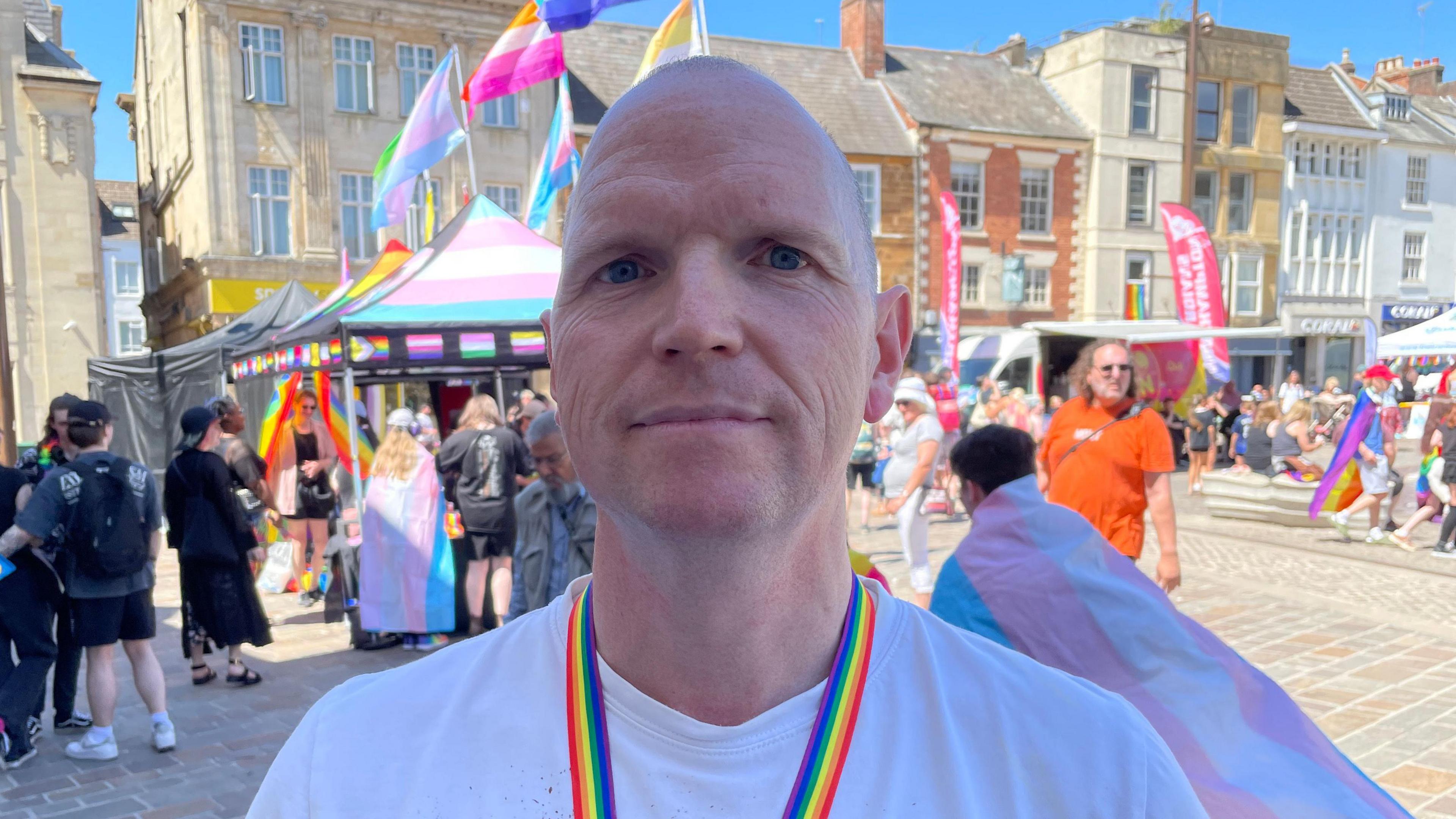 A man in a white tshirt faces the camera in a head and shoulders image. He is standing in a market square which has colourful flags and stalls around it.