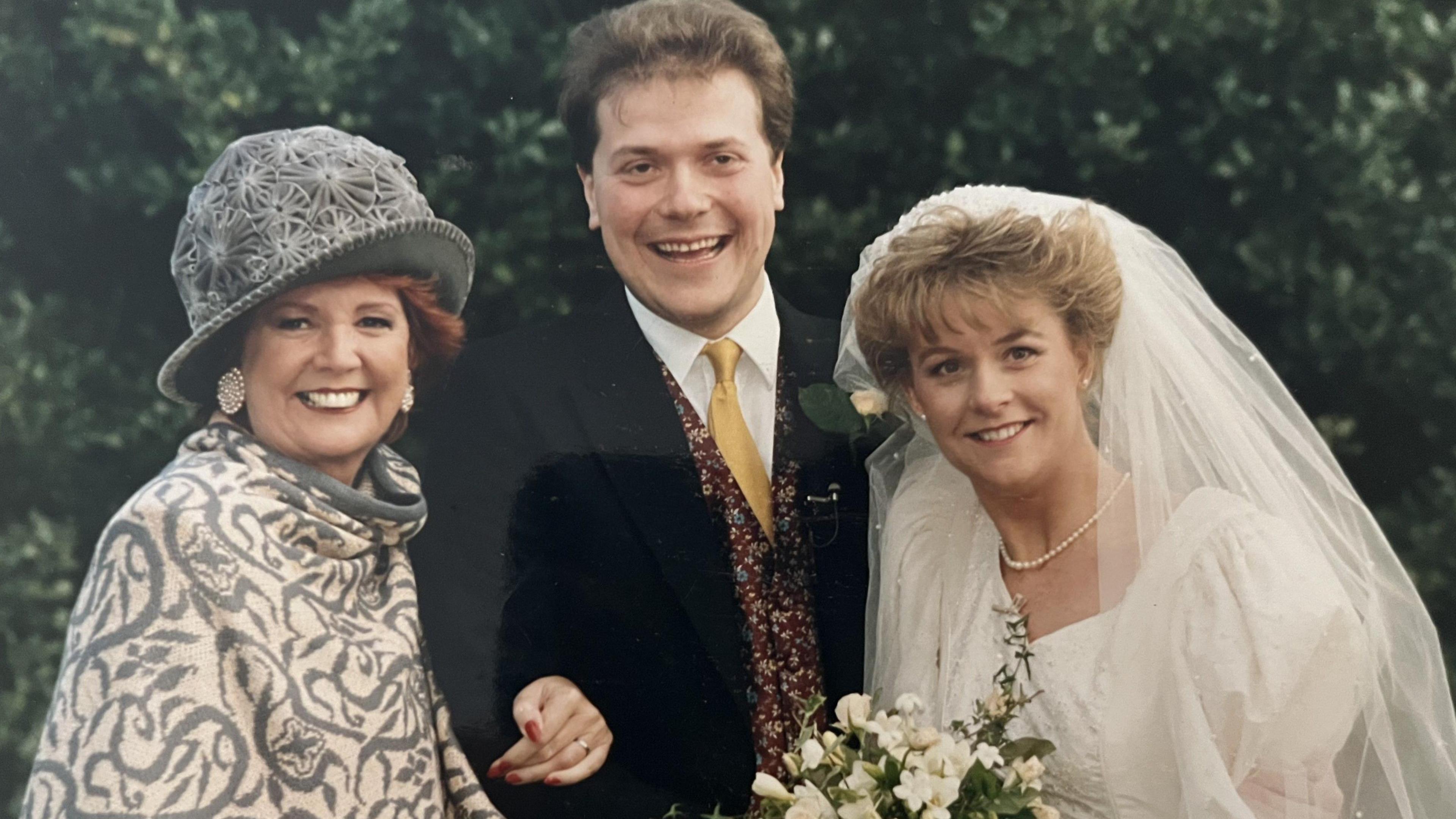 A man in a suit with yellow tie, brown hair and a red patterned waistcoat smiles at the camera, to the right is a woman with blonde hair who also smiles at the camera, wedding veil pushed back and in a wedding dress and pearl necklace. She holds white flowers. To the left is Cilla Black, with a blue and white patterned poncho, hat and big oval earrings.