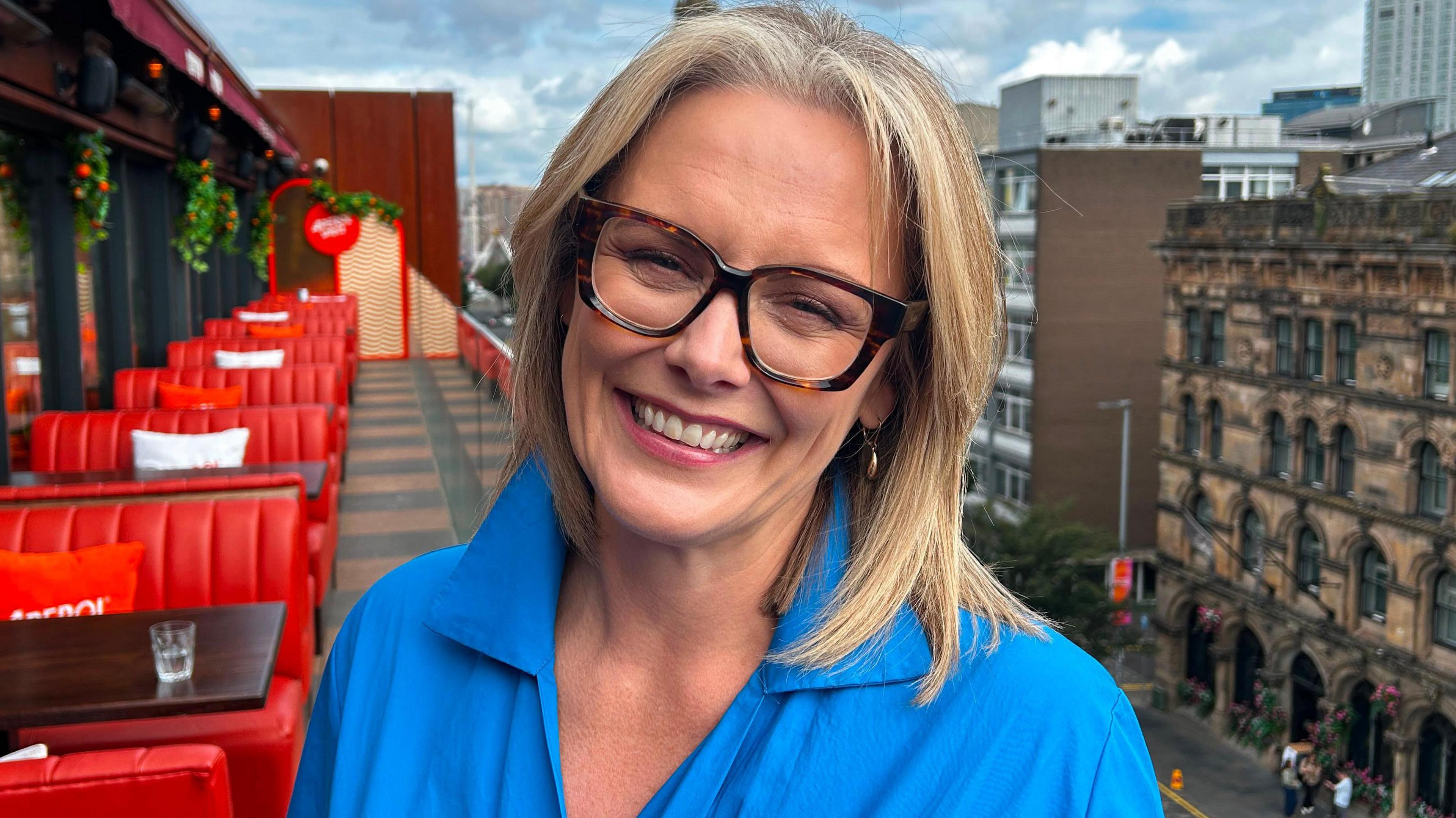 A woman in a blue shirt with blonde hair and glasses. She is standing on the balcony of a building with red sofas behind her. The sky is blue with grey clouds.