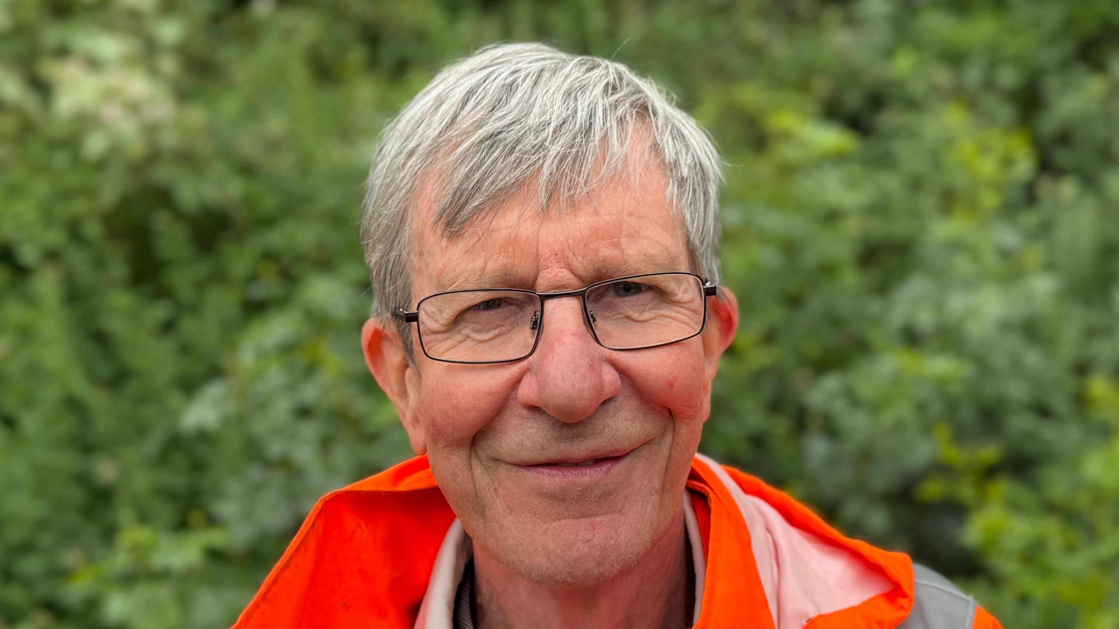 A man wearing an orange high visibility jacket in front of a hedge. He is smiling straight at the camera and wearing glasses.
