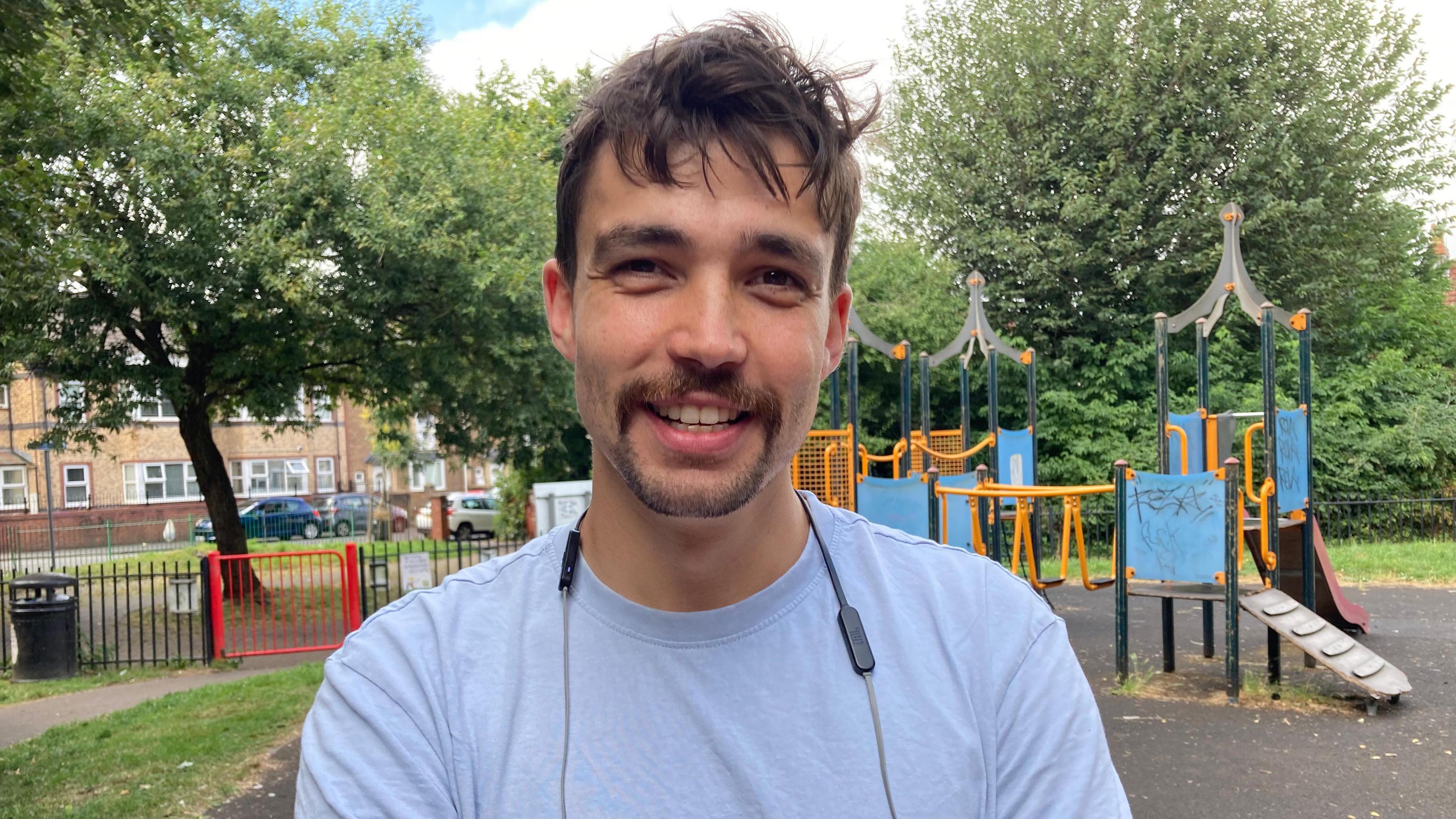 Matt Hollinshead, pictures smiling in front of a playground. He has dark hair and is wearing a light-coloured t-shirt and a pair of headphones around his neck. 