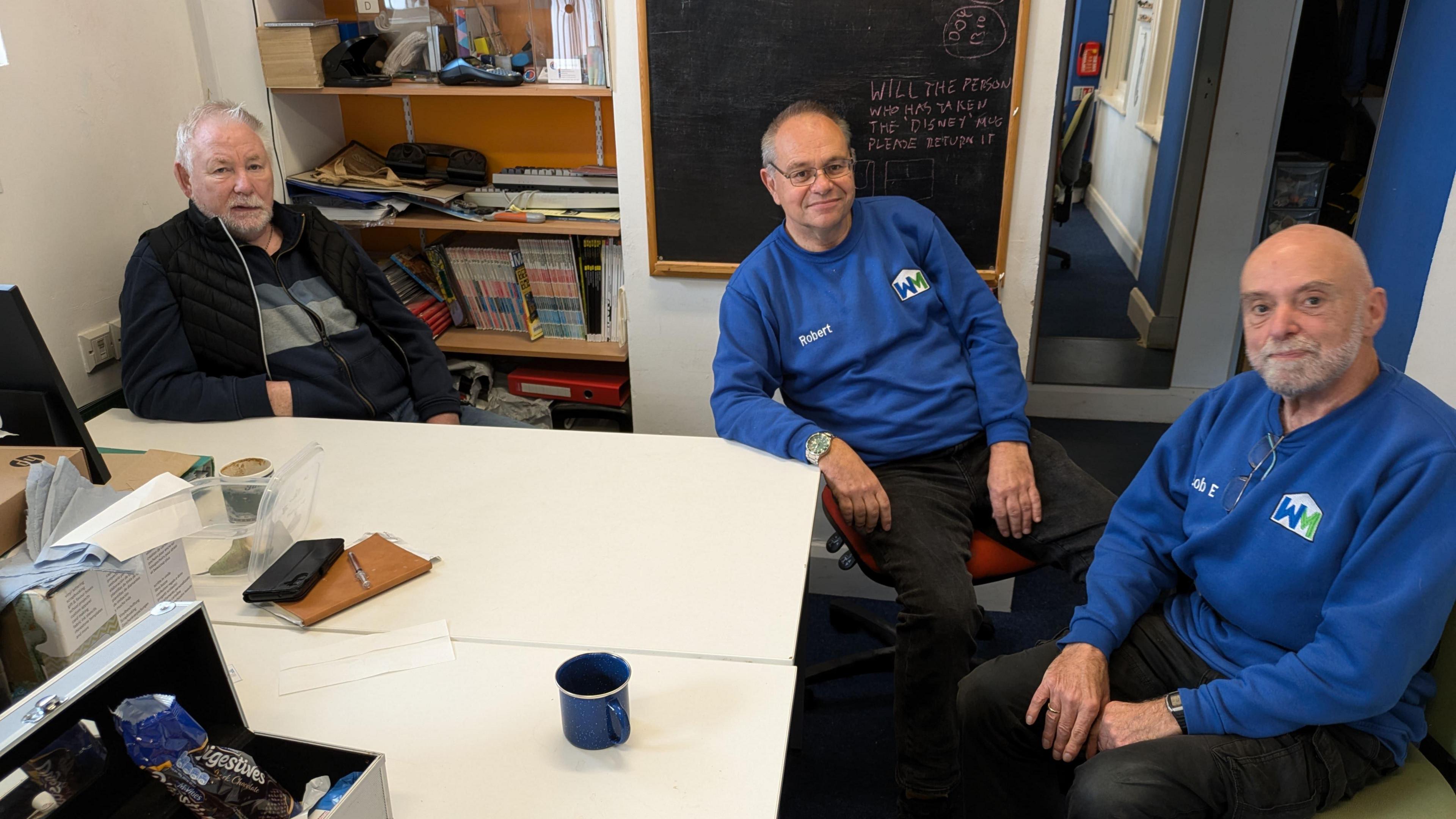 Three men sit around a white table, which has a coffee mug, a box of biscuits and other various items on it. Two are wearing blue jumpers, the other a gillet on top of a navy tracksuit.