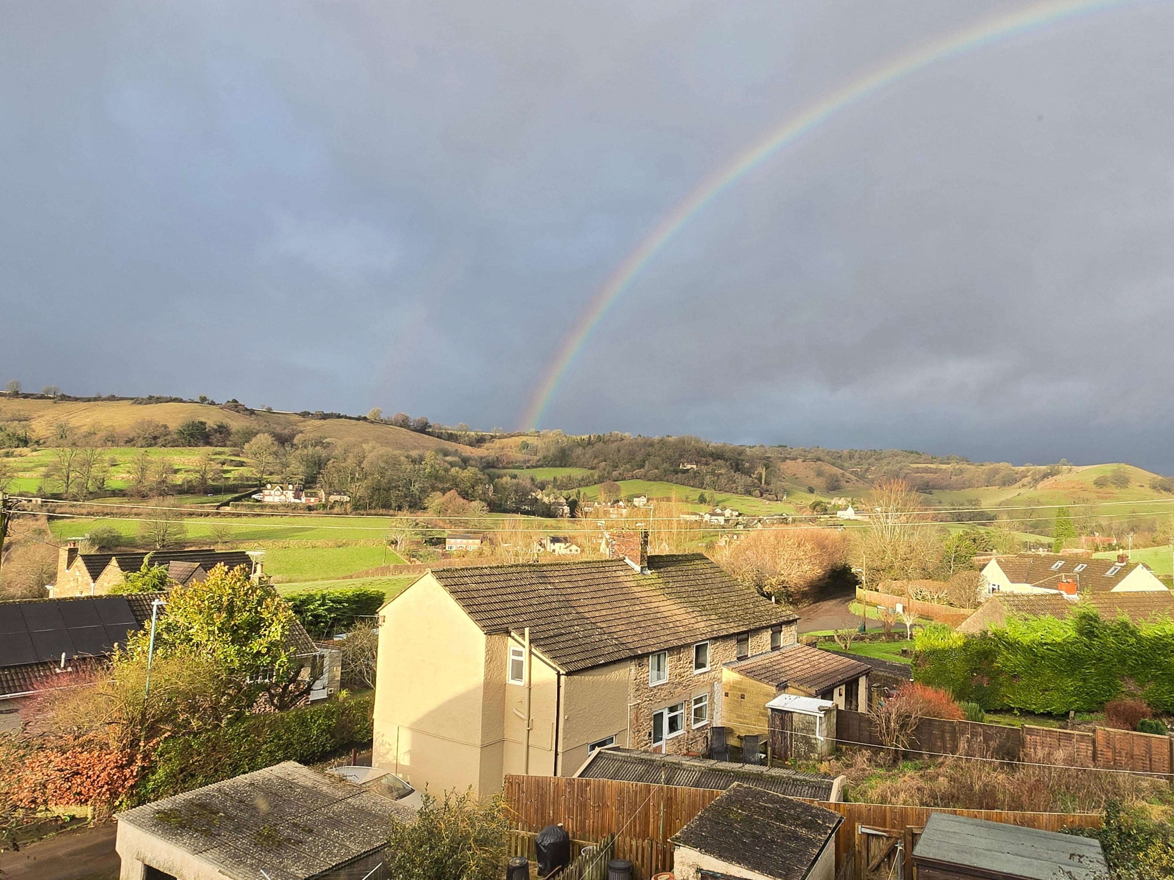 A photo of a row of three houses pictured in front of green hills. The sky is grey and a rainbow appears to be shooting out of the centre of the hills. 