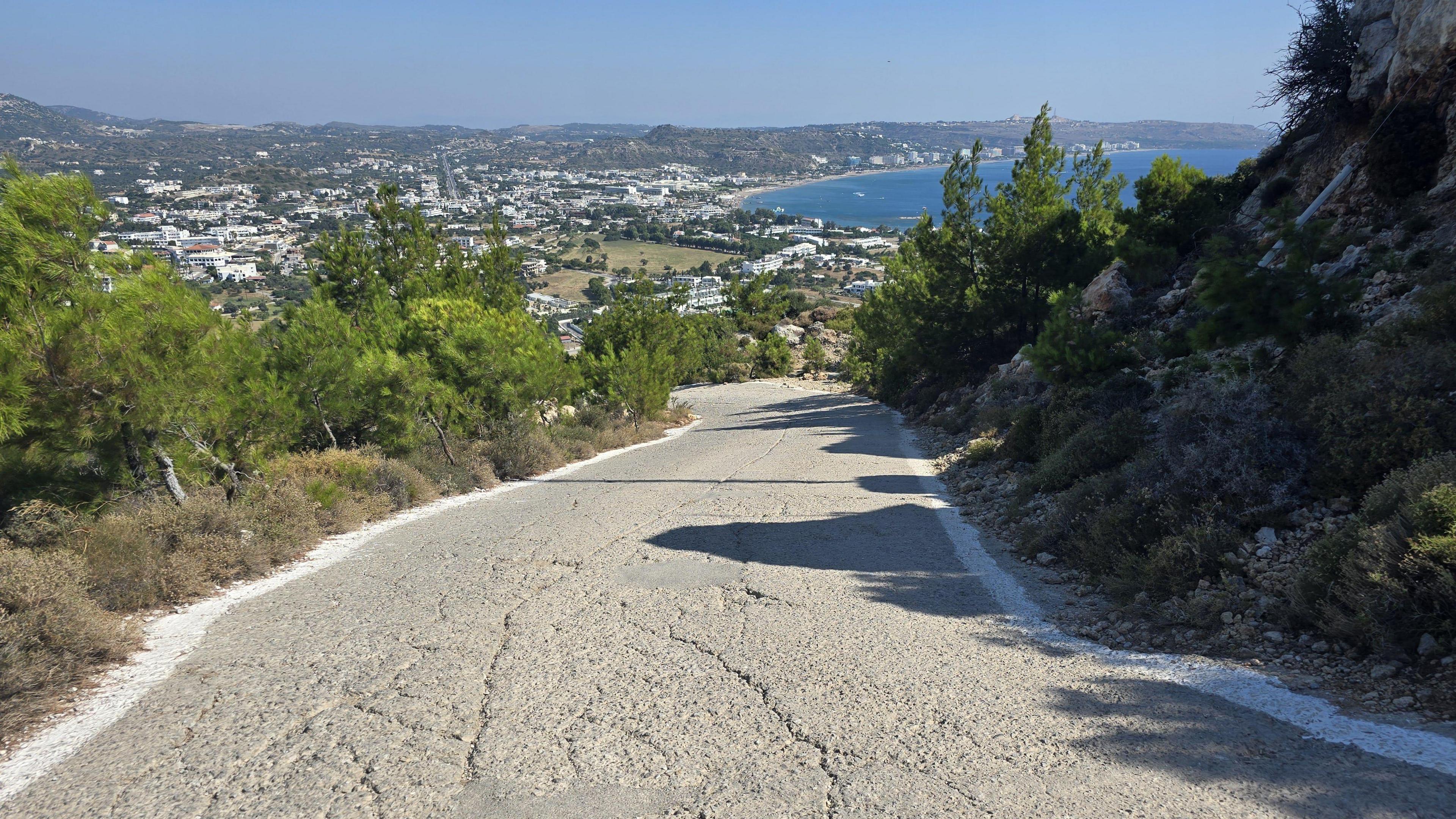 An empty hill road in a sunny place. Shrubs flank the road on both sides. The sea can be seen in the far distance.