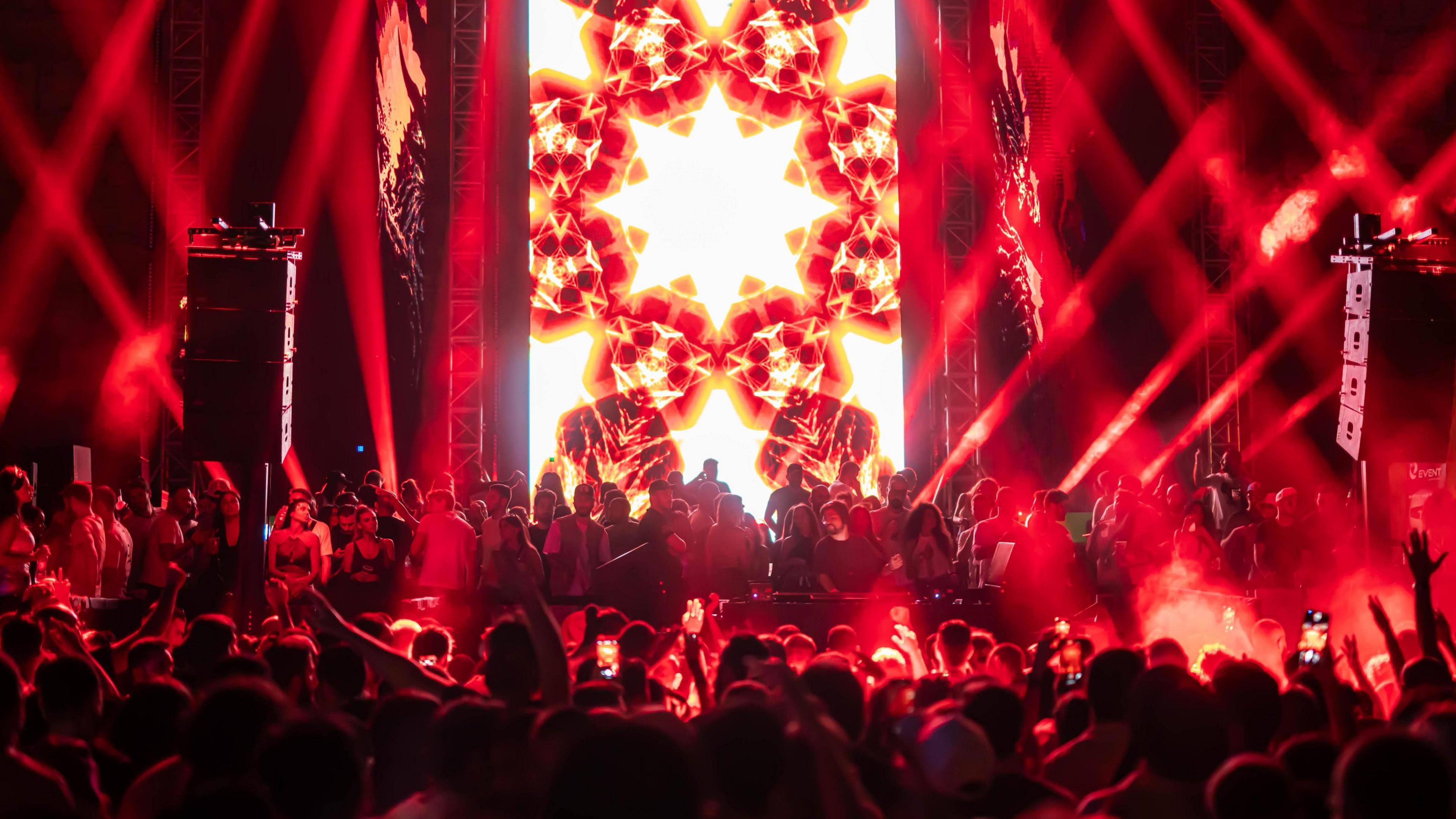 Red, black and white lights glow behind a DJ and a crowd on and in front of a the stage at an electronic music concert in Syria 