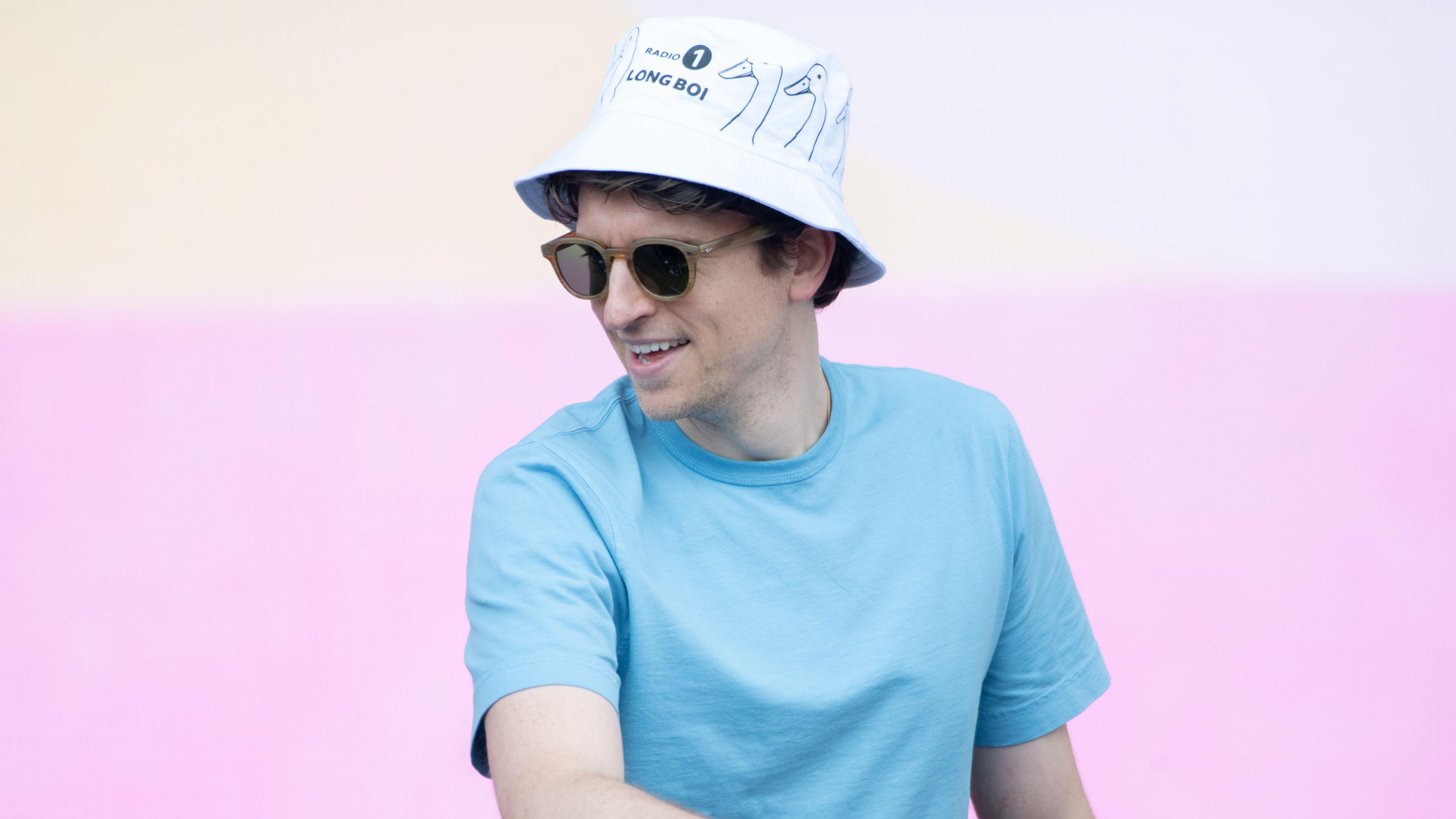 Greg James DJing at Radio 1's Big Weekend in Luton in 2024. He's wearing a white bucket hat, which has the words "Long Boi" written on it, along with three black and white drawings of ducks. He is wearing sunglasses, and a blue t-shirt.