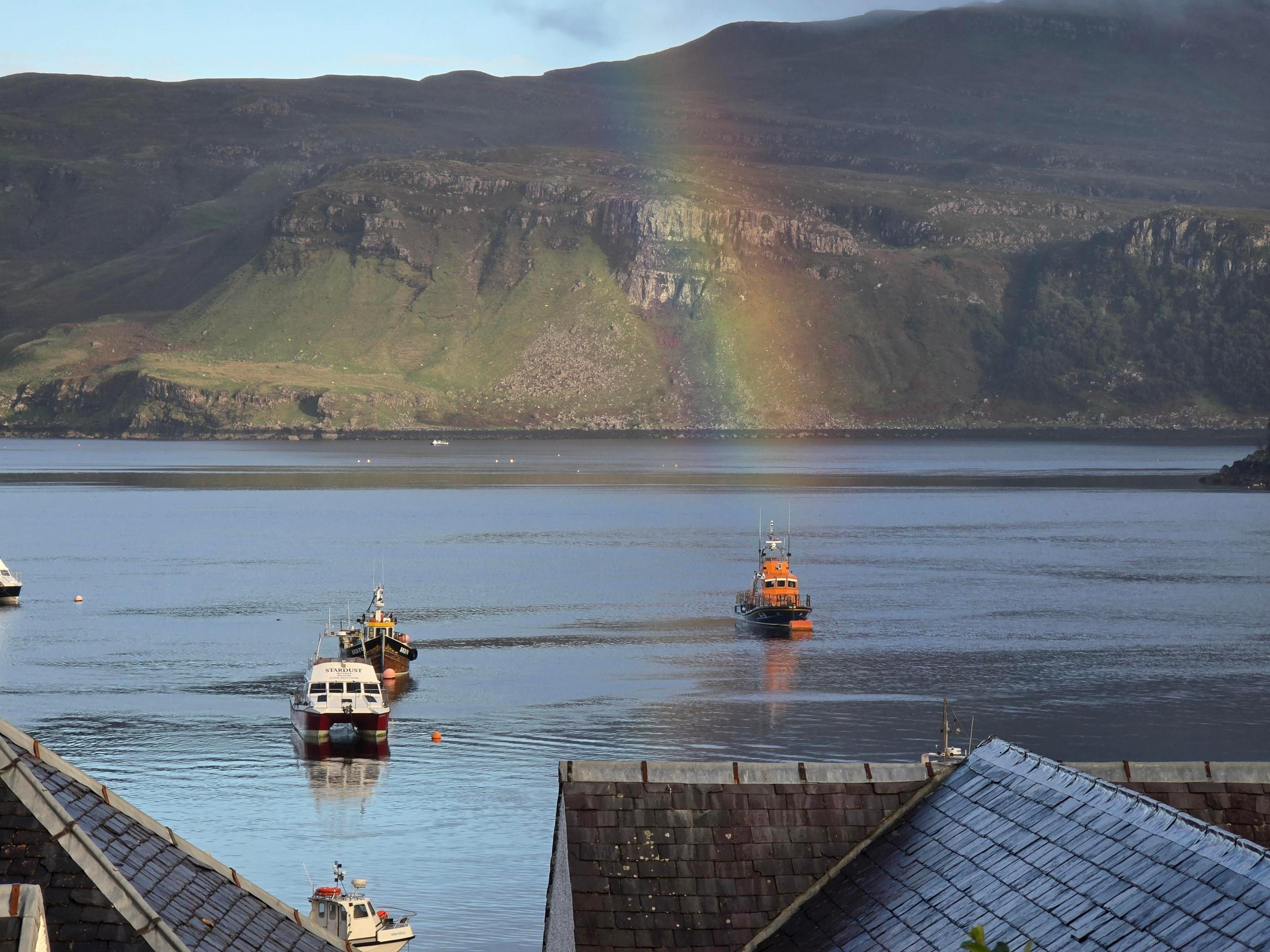 A view over some rooftops of a loch with several boats. A rainbow appears to stretch down, right over a lifeboat.