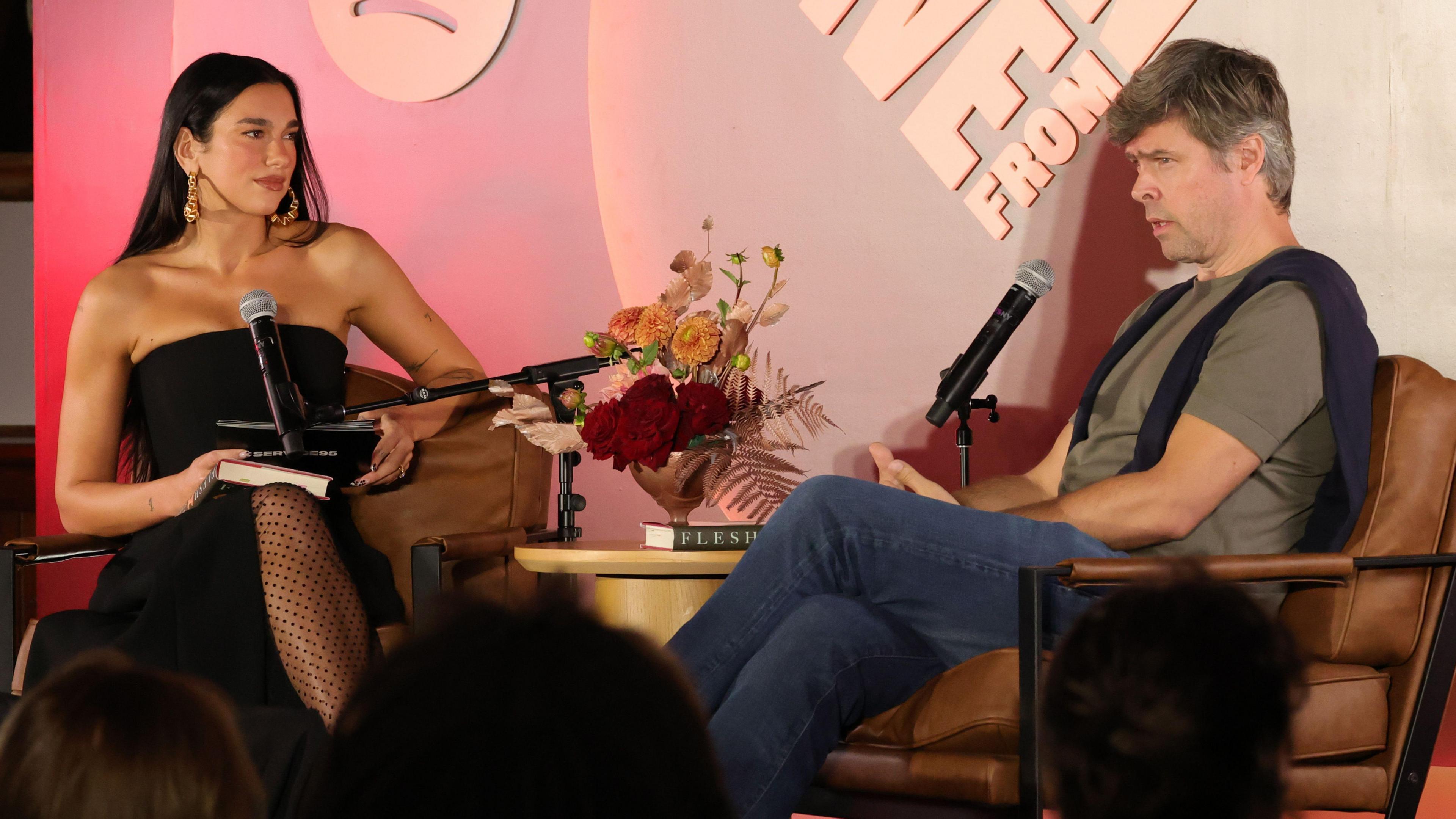 Dua Lipa and David Szalay sitting on a stage in front of microphones