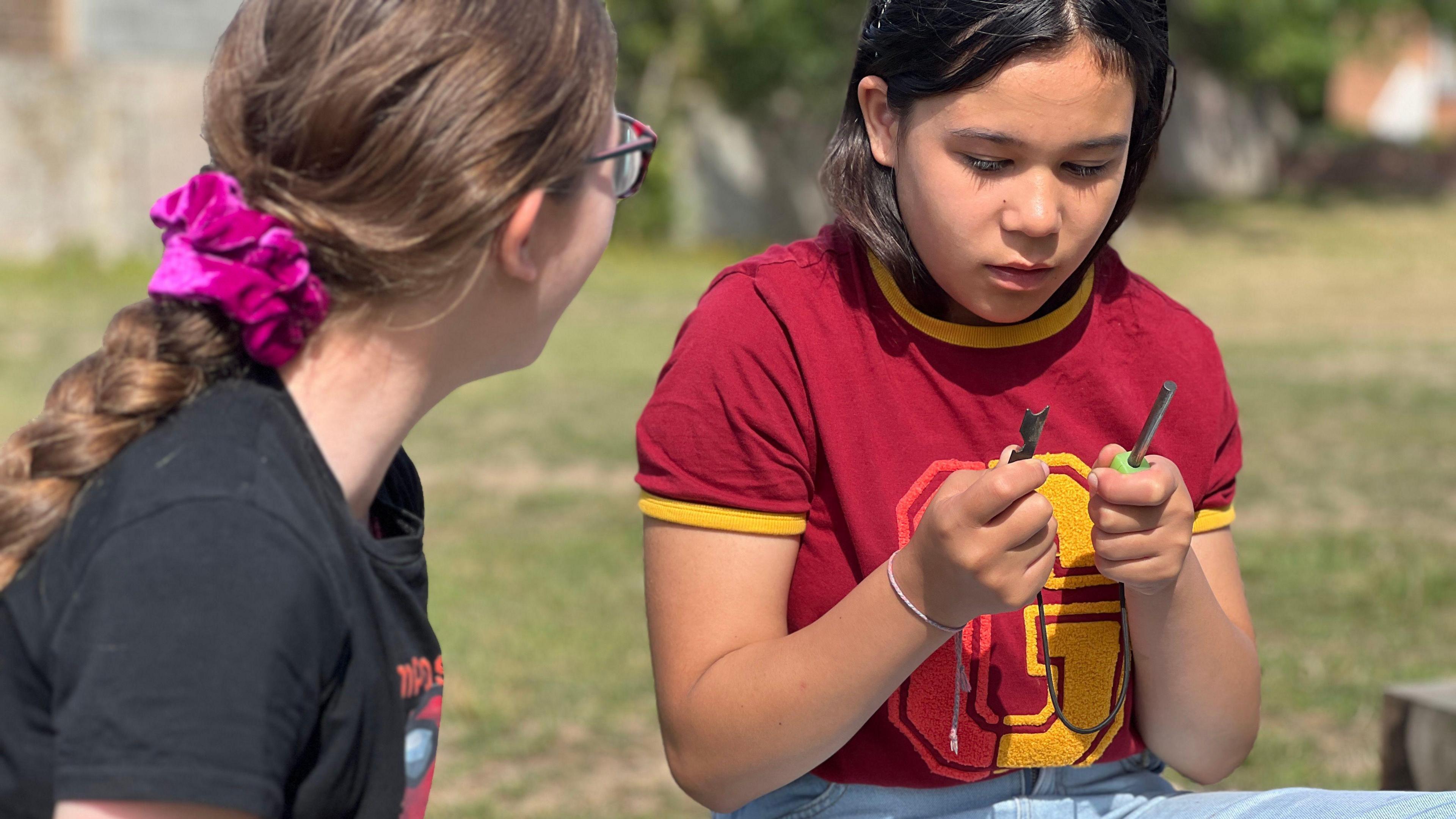 Two girls sit next to each other as one learns how to start a fire