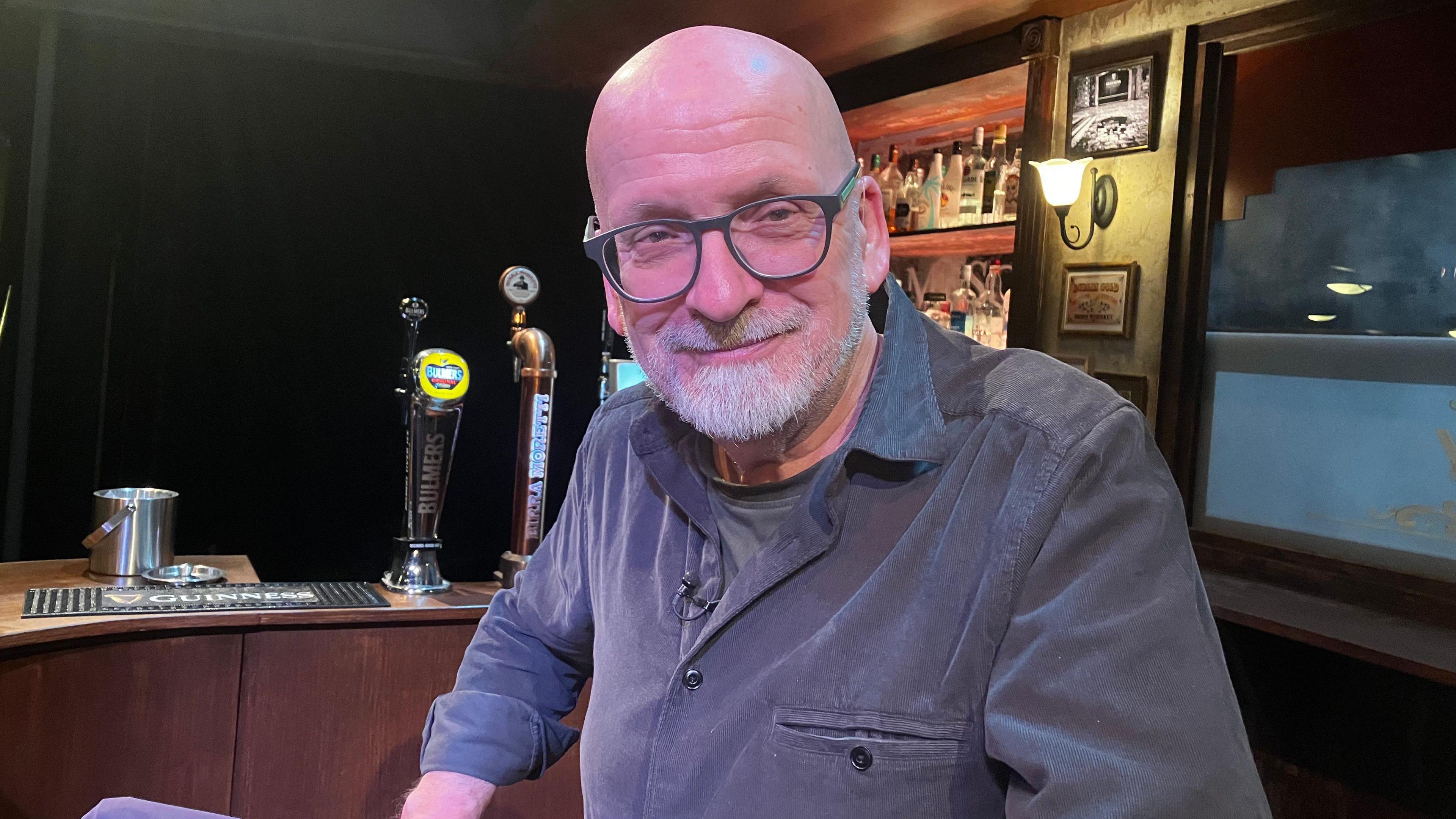 A man with a bald head, glasses and a white beard sits in a bar on a theatre stage. He is wearing a grey t-shirt under a blue-grey shirt.
