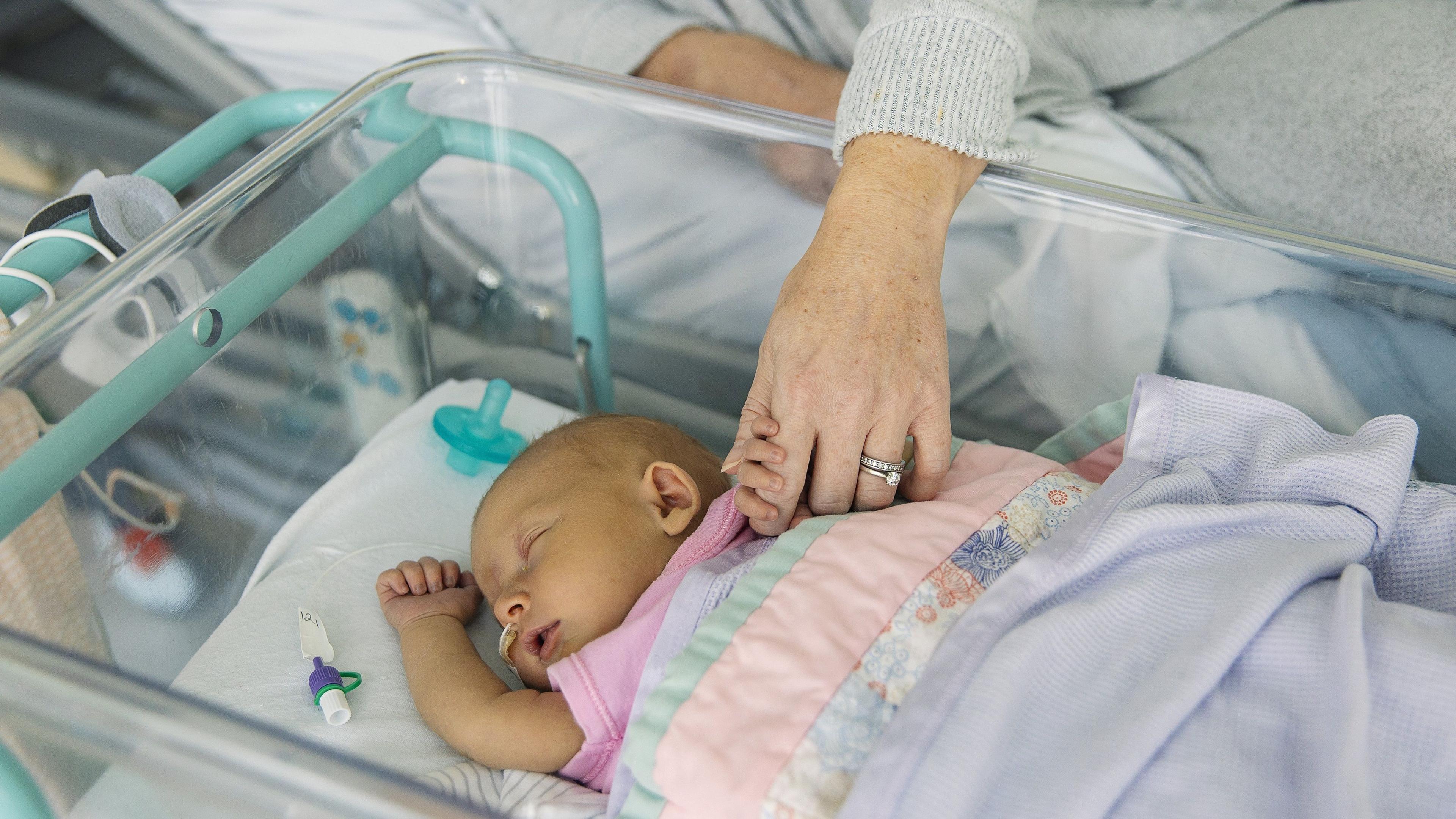A newborn baby sleeps in a bassinet while its mother, lying in a hospital bed, holds one of its hands.