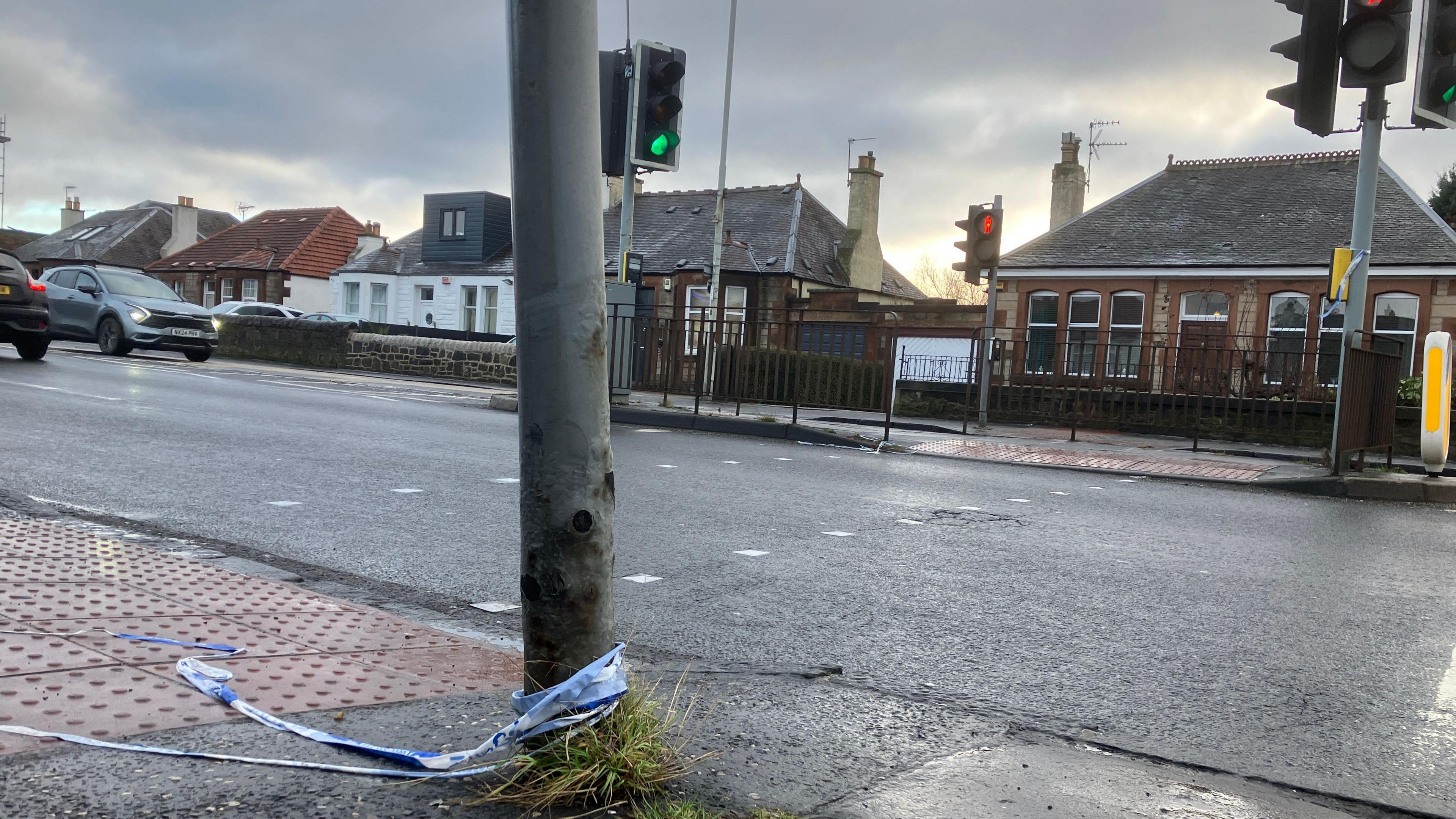 Road with cars on it with houses down either side. There is a pedestrian crossing with police tape attached to it.