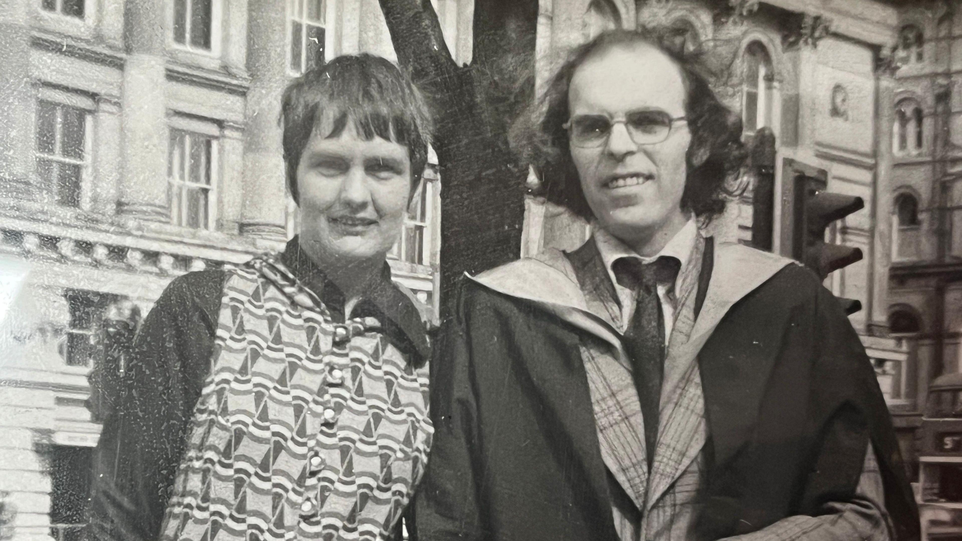 Black and white photograph from the 1970s of the Wetherells. Pat is standing to the left of the shot and has short dark hair and a patterned top with plain sleeves, in the style of the times. Ivan has receding but otherwise long dark hair and is wearing spectacles, a graduation gown over a checked jacket, light shirt and dark tie.