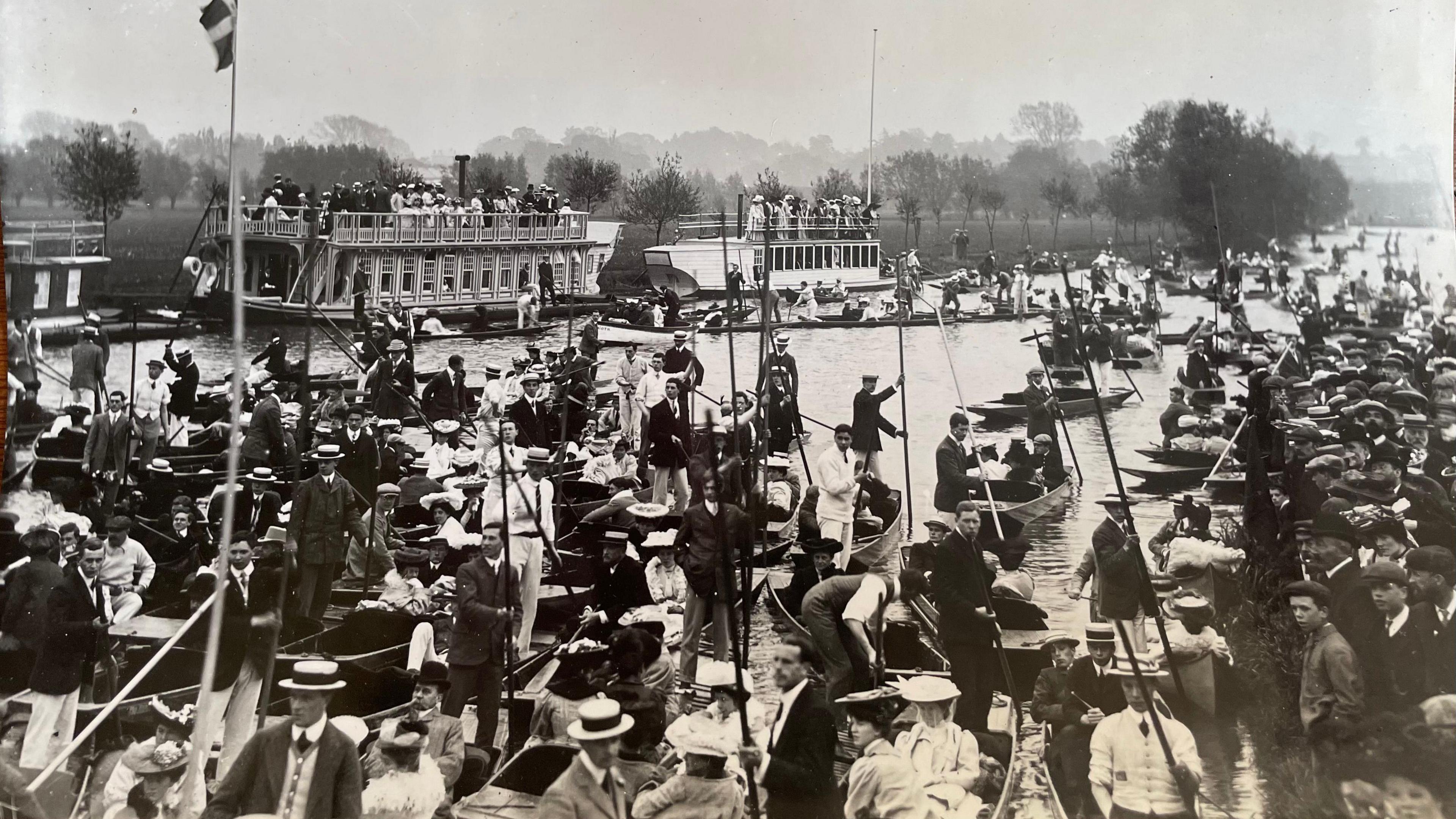 A black and white archive image of people on boats at the river in Oxford and three college barges lined by the bank.