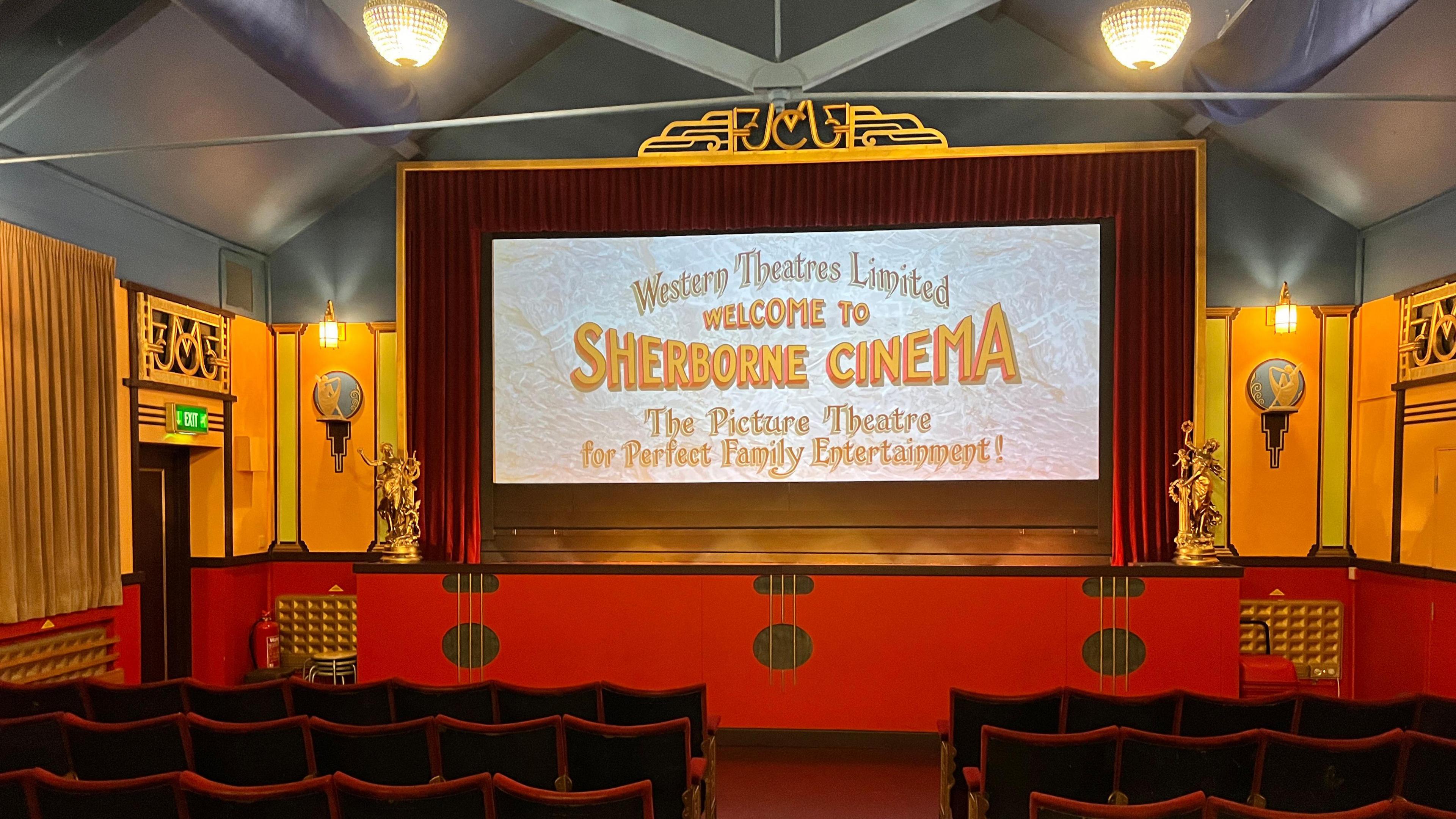 The auditorium of the small Sherborne Cinema has a small number of rows of deep red fold-down seats, which face a one-metre high stage. Above that is the screen which is surrounded by a red curtain. The walls are painted deep orange and bright yellow, and there are golden statues on either side of the screen.