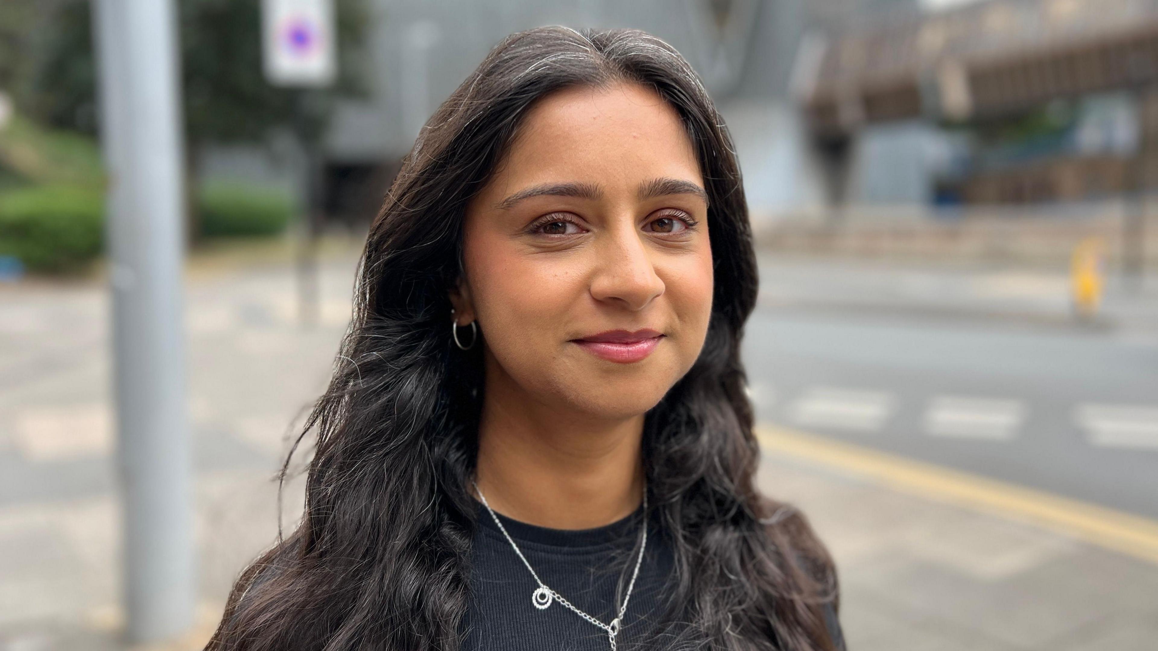 Kauser Husain looks at the camera. She is wearing a black T-shirt and silver necklace. She has long brown hair and pink lipstick.