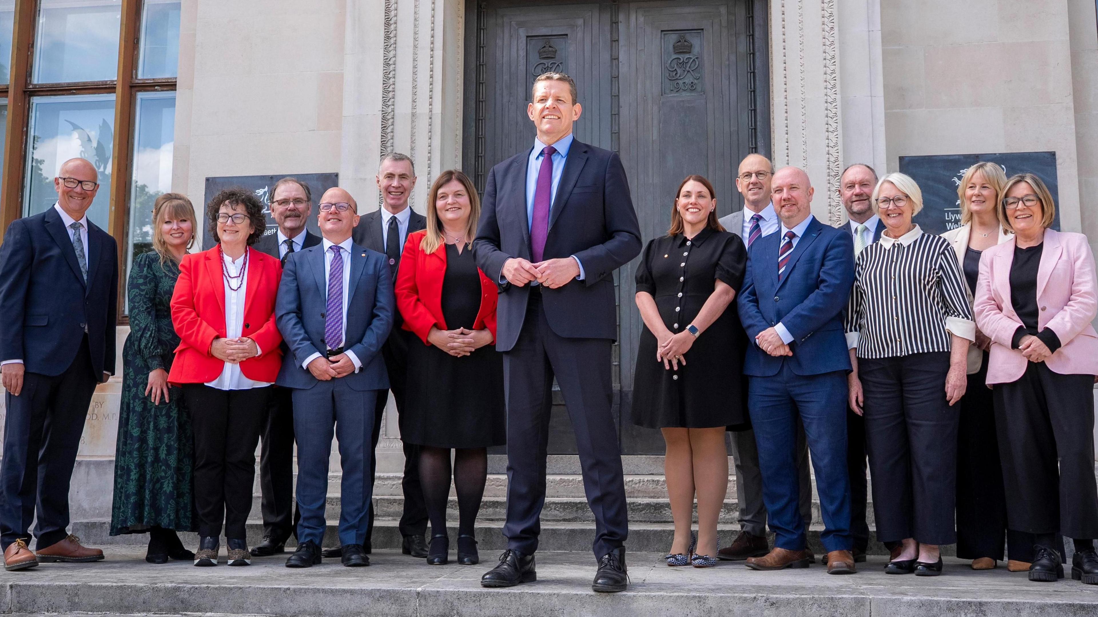 Mae'r llun yn dangos Rhun ap Iorwerth yn sefyll o flaen aelodau ei gabinet ar risiau pencadlys Llywodraeth Cymru. Mae'n ddiwrnod braf ac mae pawb yn gwenu.