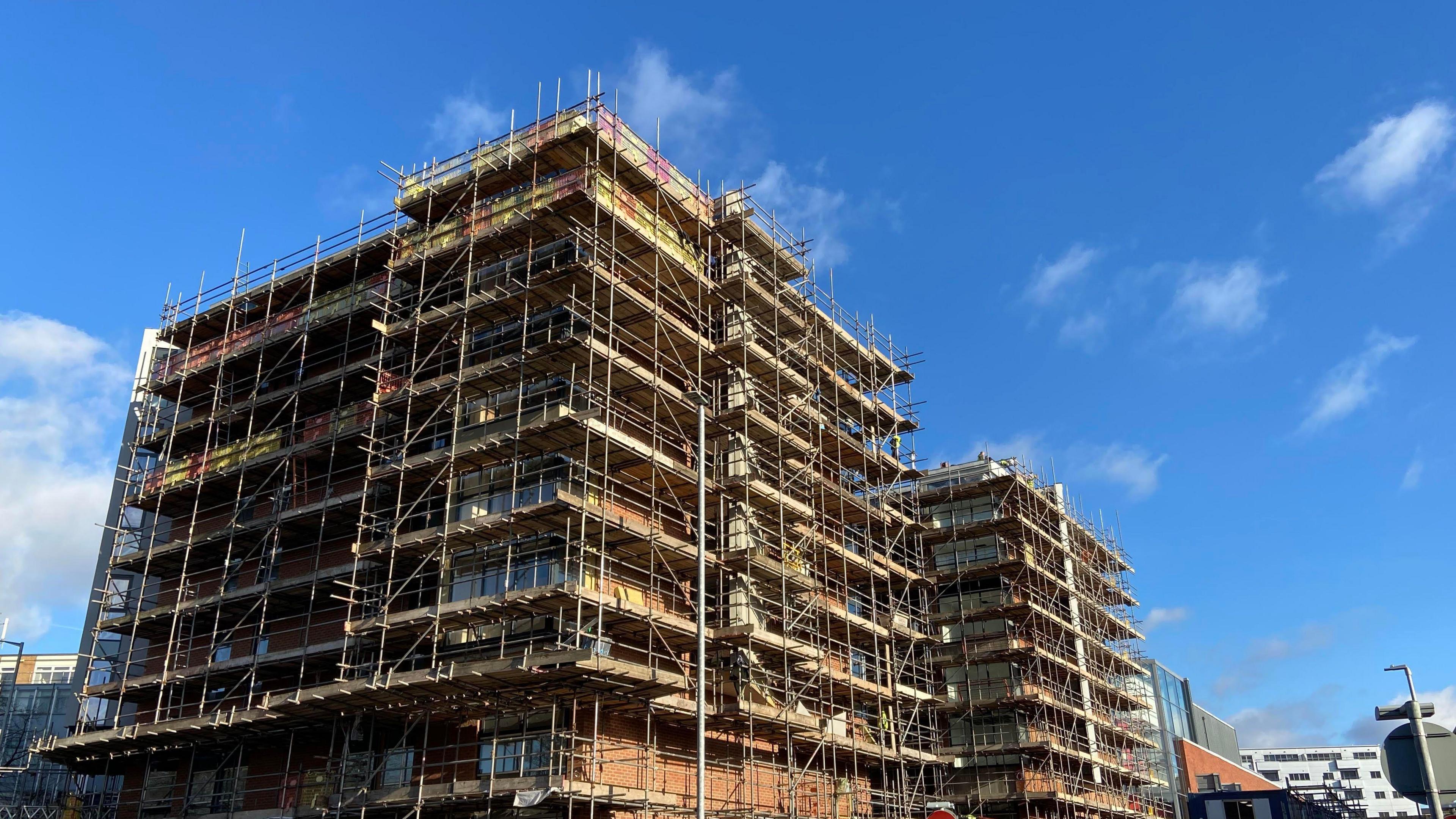 A seven-storey building, is surrounded by scaffolding, can be seen against a blue sky.