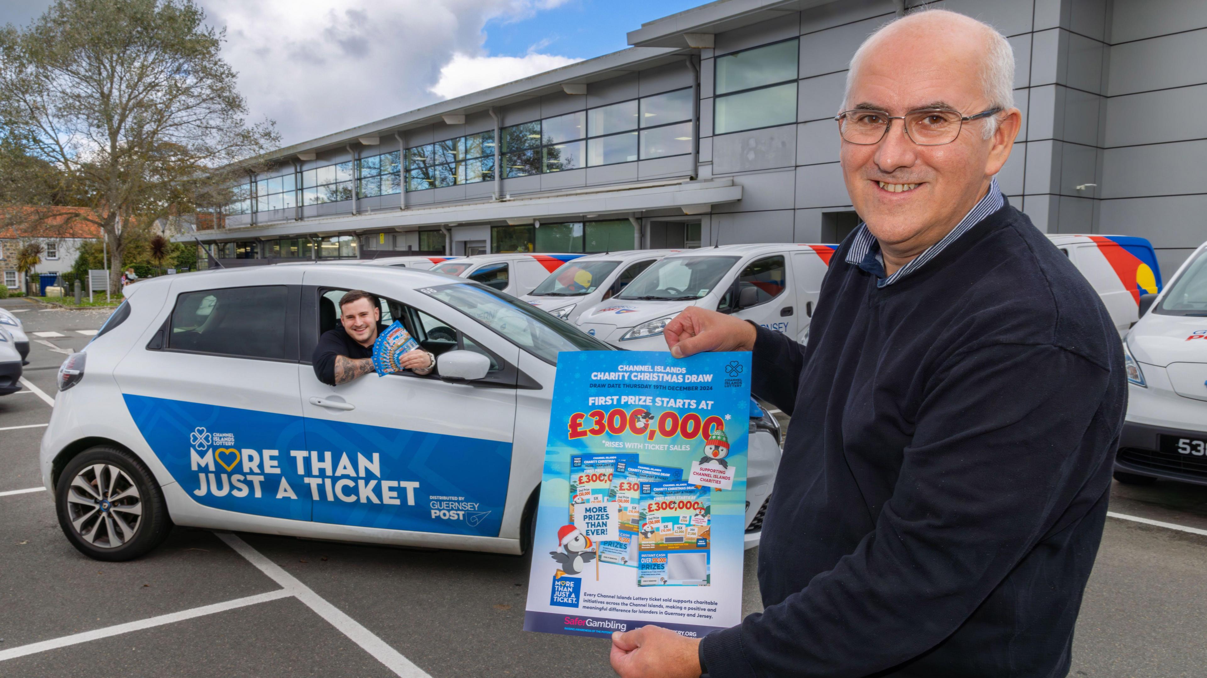 Bill Can smiling on the right side of the photograph holding a blue and white festive lottery poster which has the words 'first prize starts at £300,000'. It also has a cartoon penguin on it wearing a Christmas hat. In the background is a man leaning out of the window holding more of the posters. The car has a blue sign on the side of it which has the Channel Islands lottery logo with the words 'more than just a ticket'. Behind the car is a grey building with windows and parked vans in front of it.