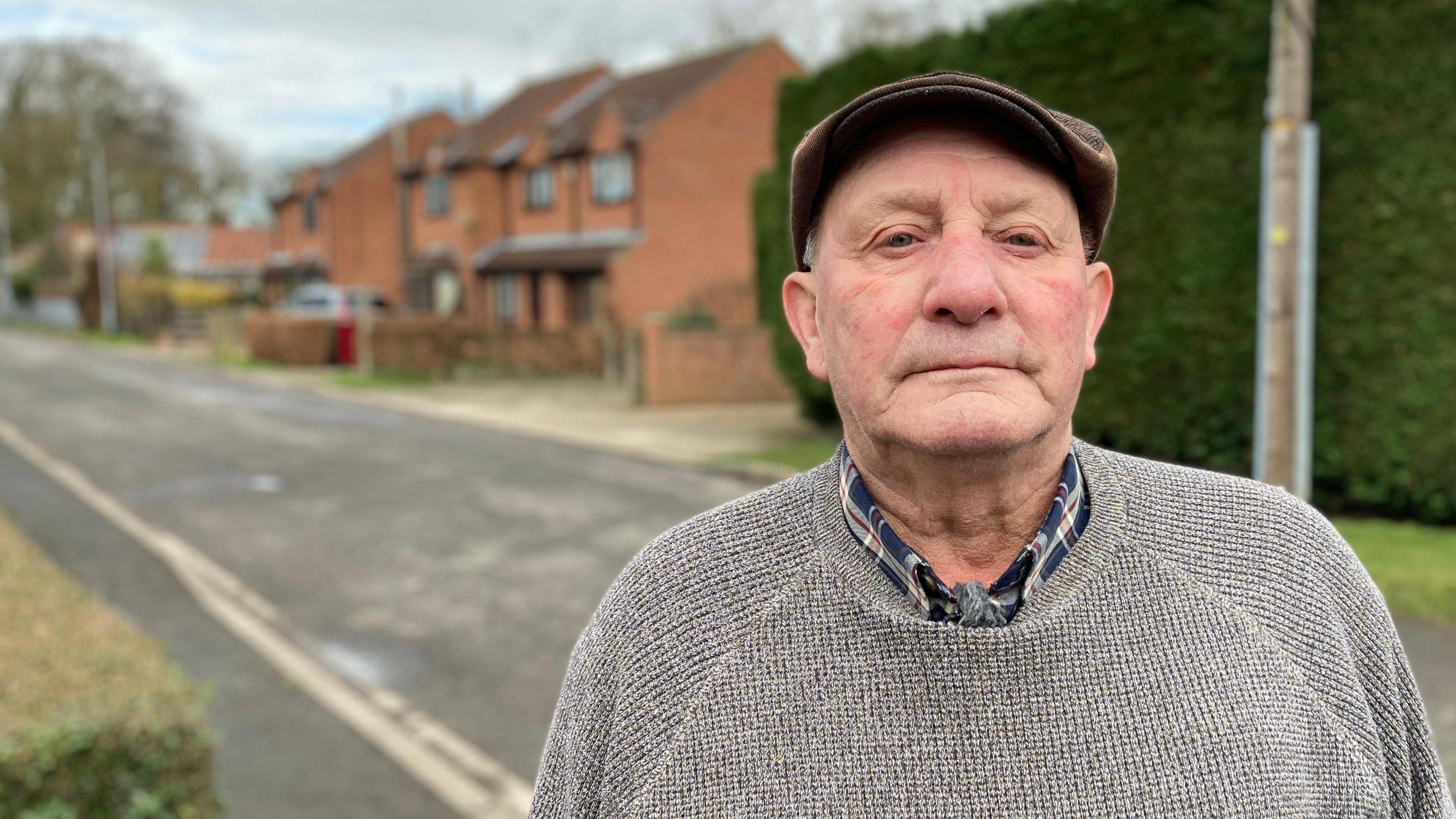 Paul is wearing a flat cap on his head and a grey knitted sweater over a blue-checked shirt.