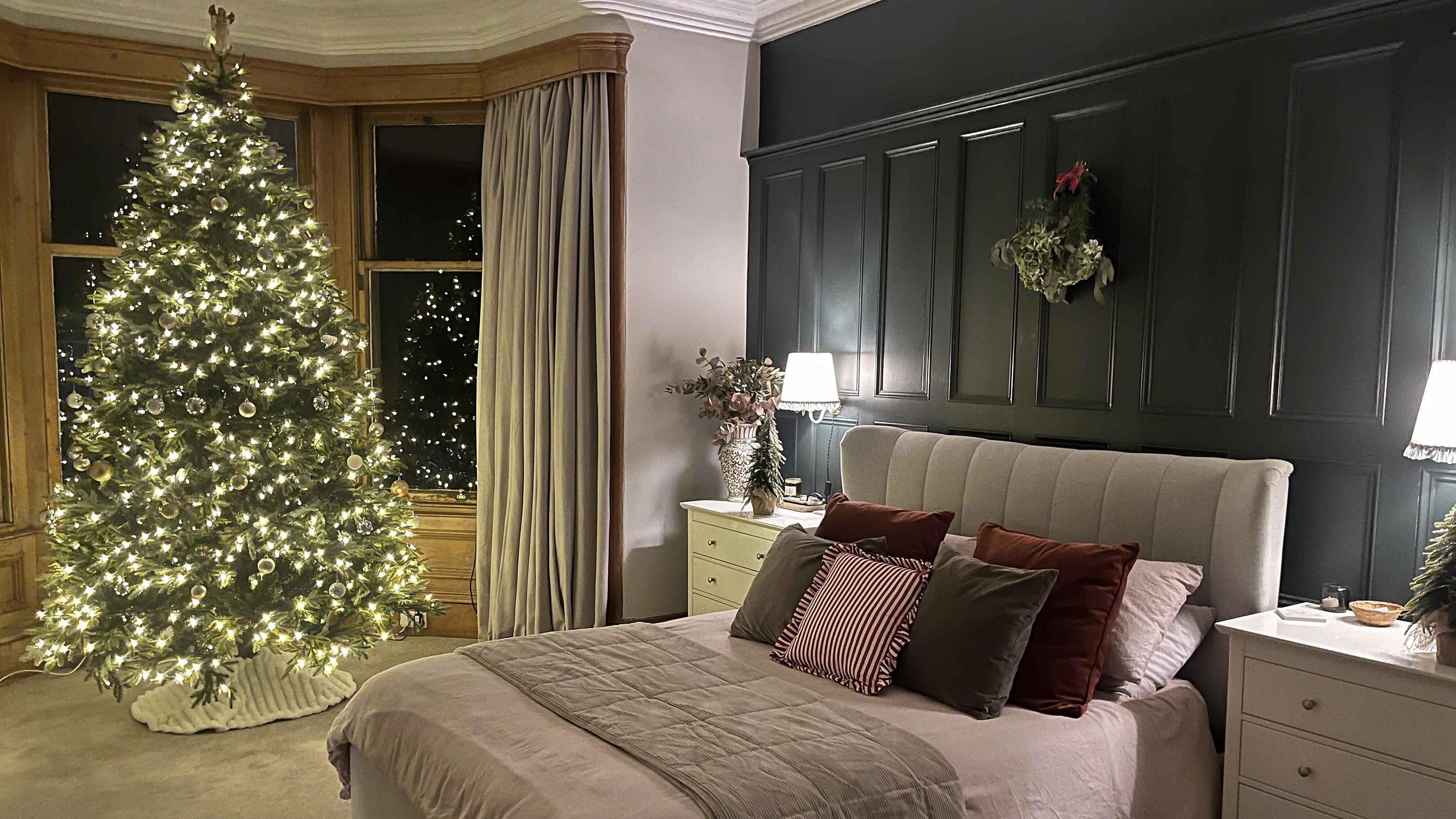 A bedroom, with a large Christmas tree in front of a bay window. The tree is covered in warm white fairy lights . A wreath hangs above the bed which is cosily decorated with cushions and a throw. 