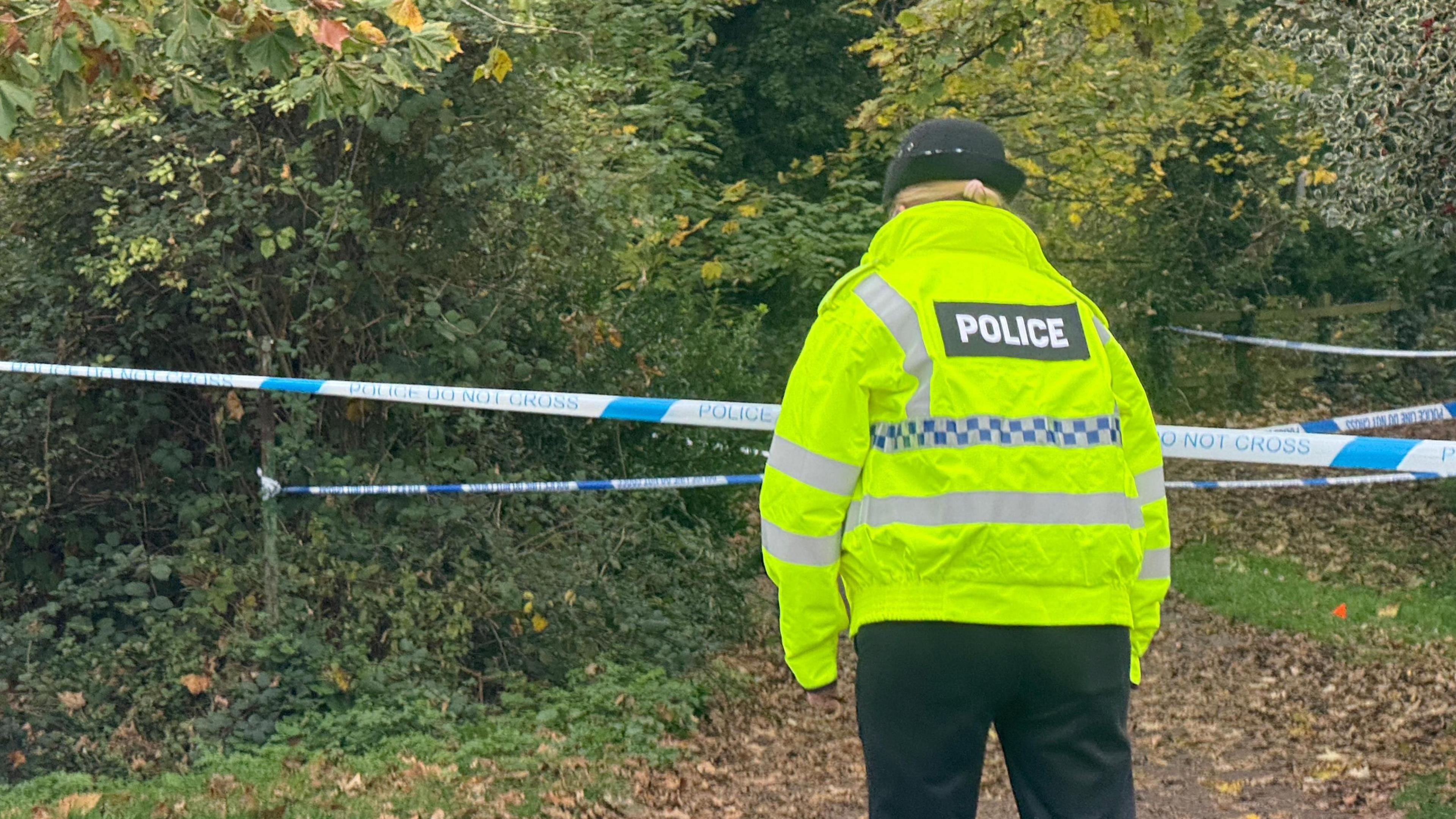 A police officer wearing a hi-vis jacket and black trousers, standing in front of blue and white police tape. The tape is cordoning off a wooded area and path with leaves on the floor.