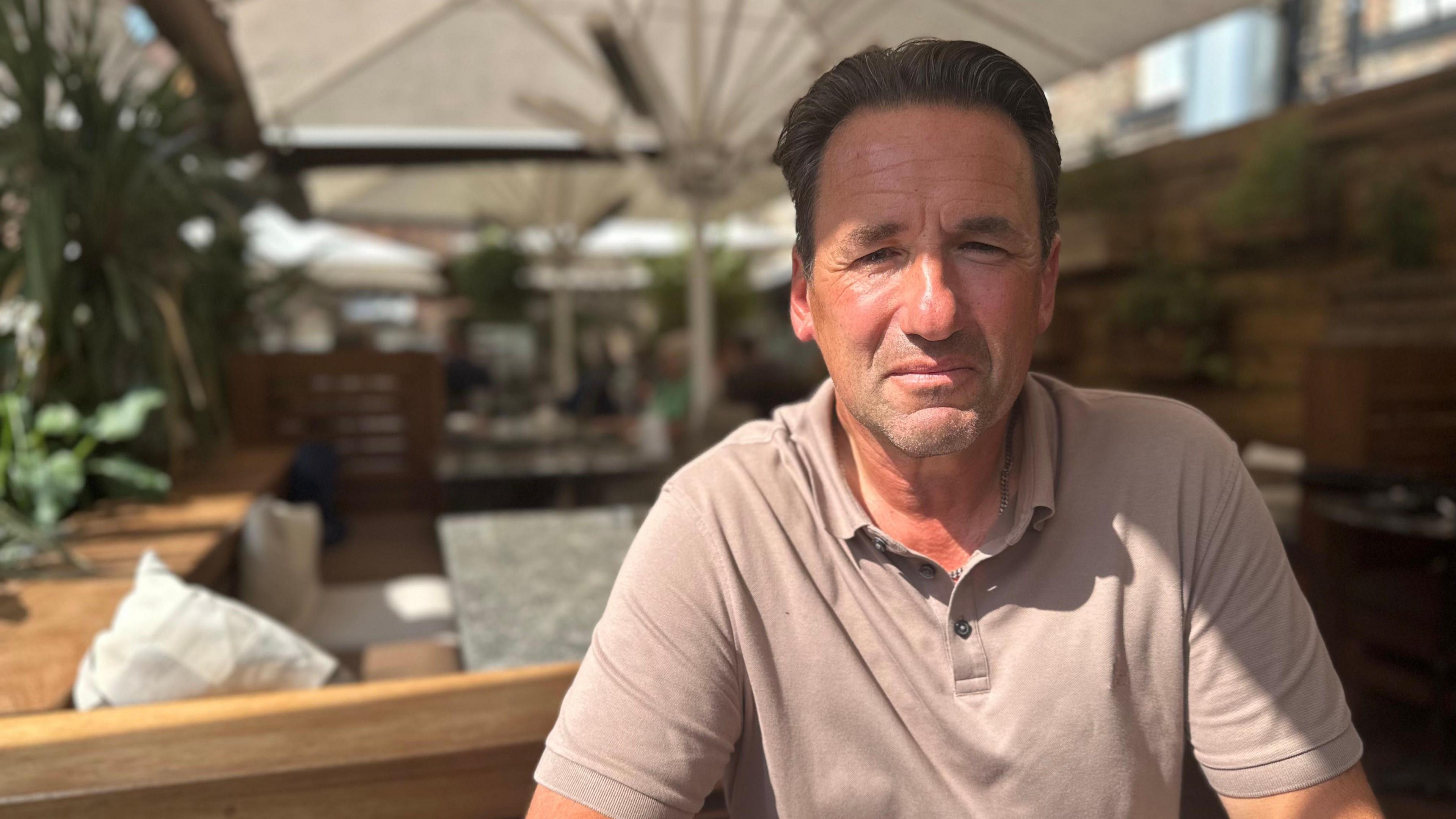 Ollie Vaulkard looking directly into the camera. He has brown hair, which is brushed back, and is wearing a light-coloured polo shirt. He is sitting in a bar's outside area which has wooden tables and benches.
